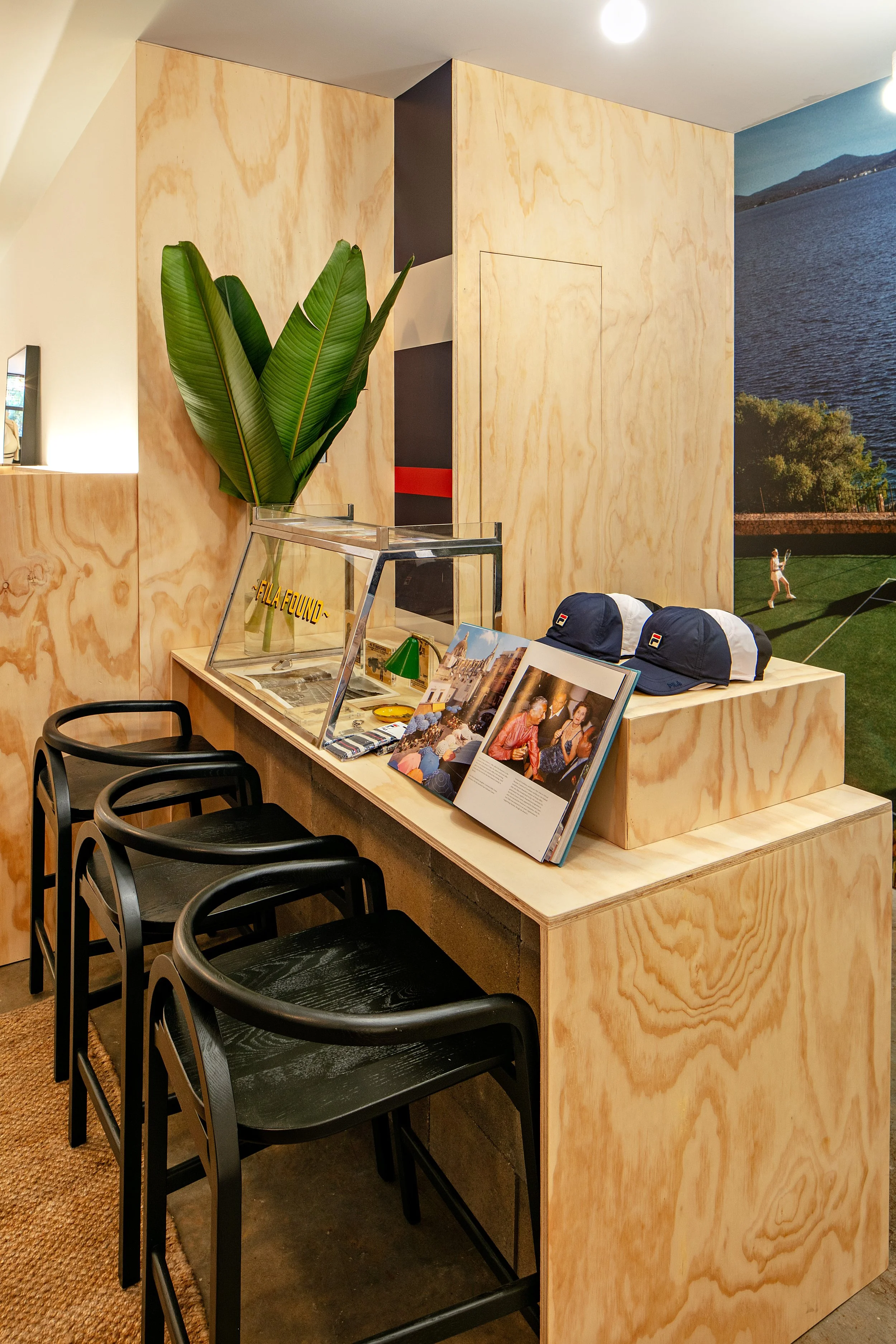 A small wooden counter in a minimalist space with black chairs lined against it. On the counter are open books and Fila caps. A glass display case with tropical leaves inside is also present. The wall is decorated with a large photo of a lake scene.