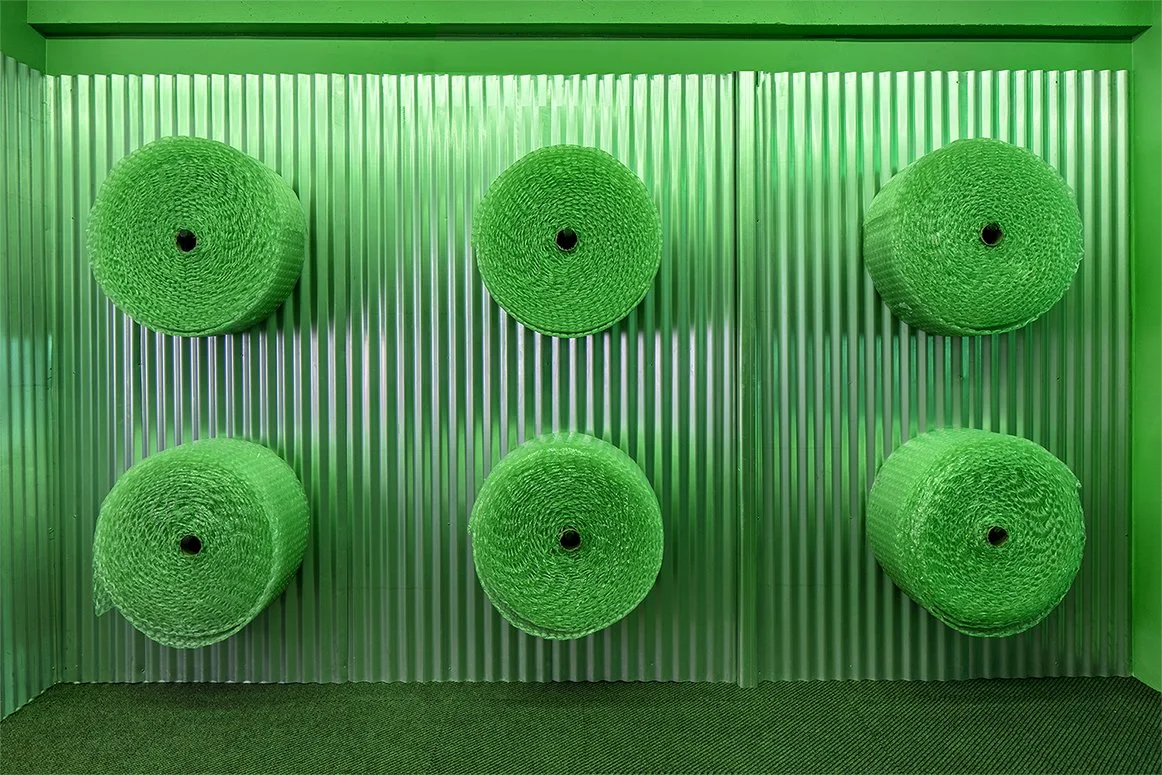 Six rolls of green bubble wrap mounted on a corrugated metal wall.