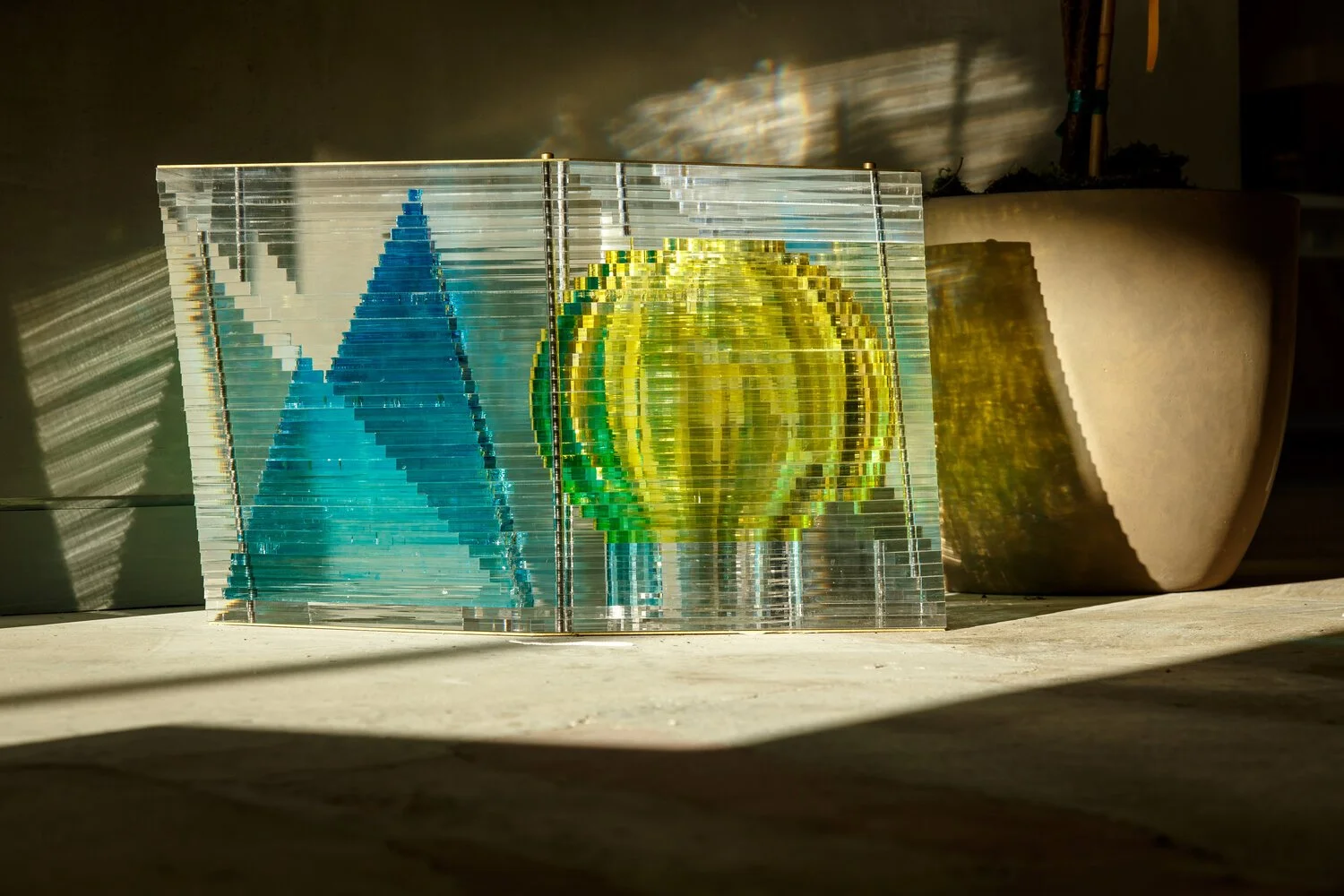 A layered glass sculpture with geometric designs, featuring a blue triangle and a yellow sphere, displayed in a sunlit room next to a large planter.