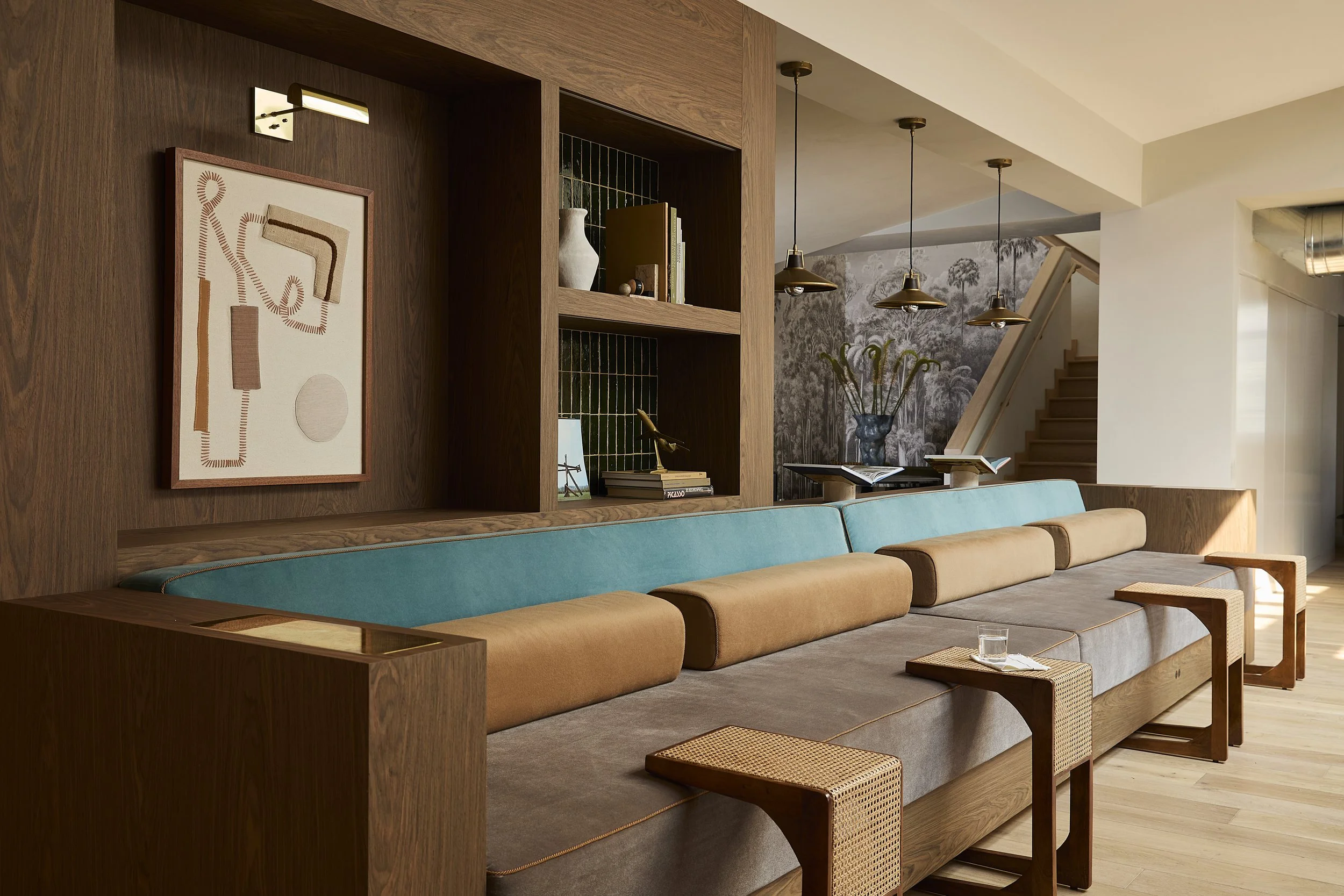 Living room with a long bench-style sofa, wooden shelving, and modern decor including a vase with flowers and books, illuminated by hanging pendant lights.