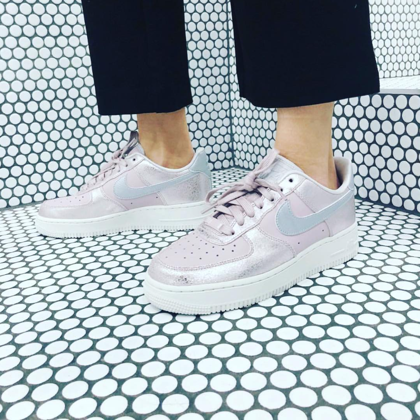 Person wearing light pink Nike sneakers with a white sole, standing on a black and white tile floor with a circular pattern.