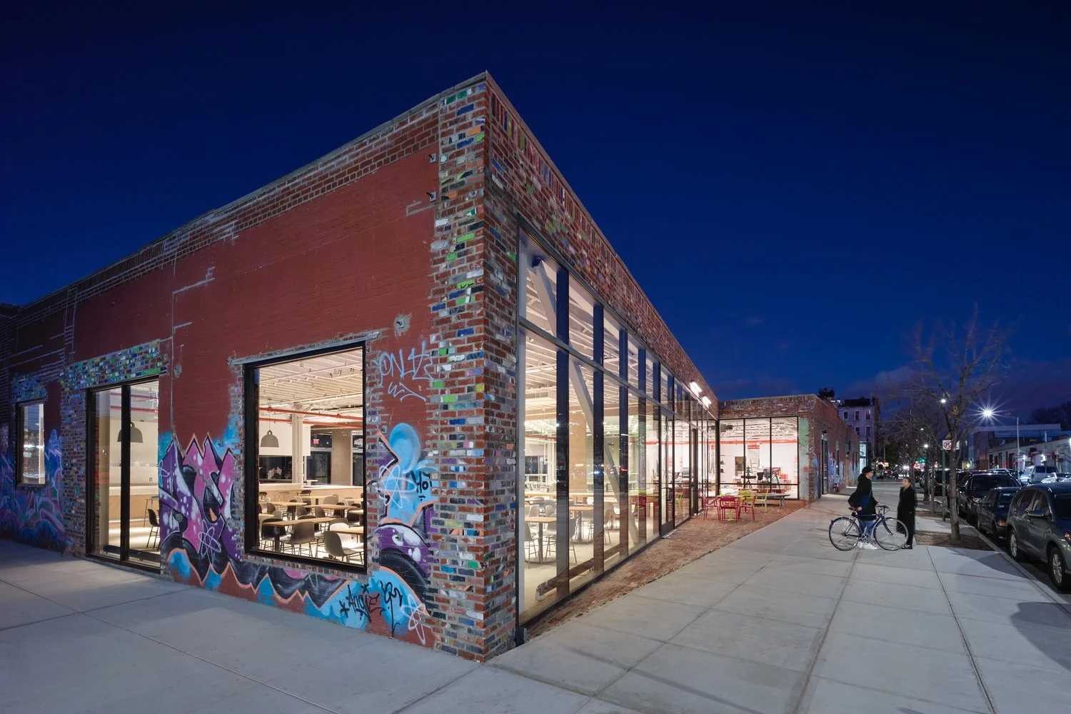 Modern brick building with graffiti at night, large windows, and interior lights visible, located on a city street.
