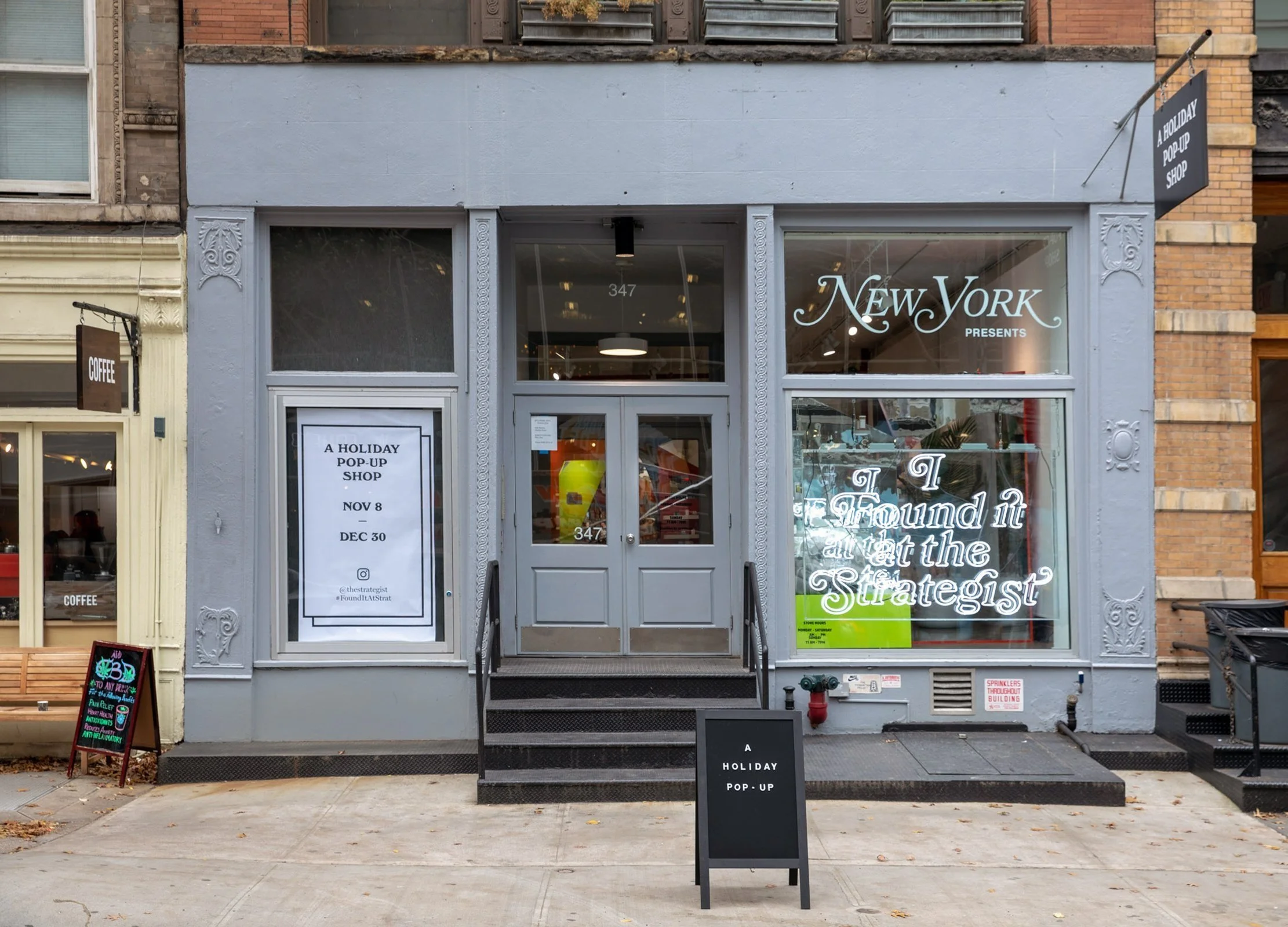 Front view of a holiday pop-up shop in New York, featuring a neon sign and promotional posters in the windows. The sign reads "I Found it at the Strategist." The sidewalk displays additional signage advertising the event.