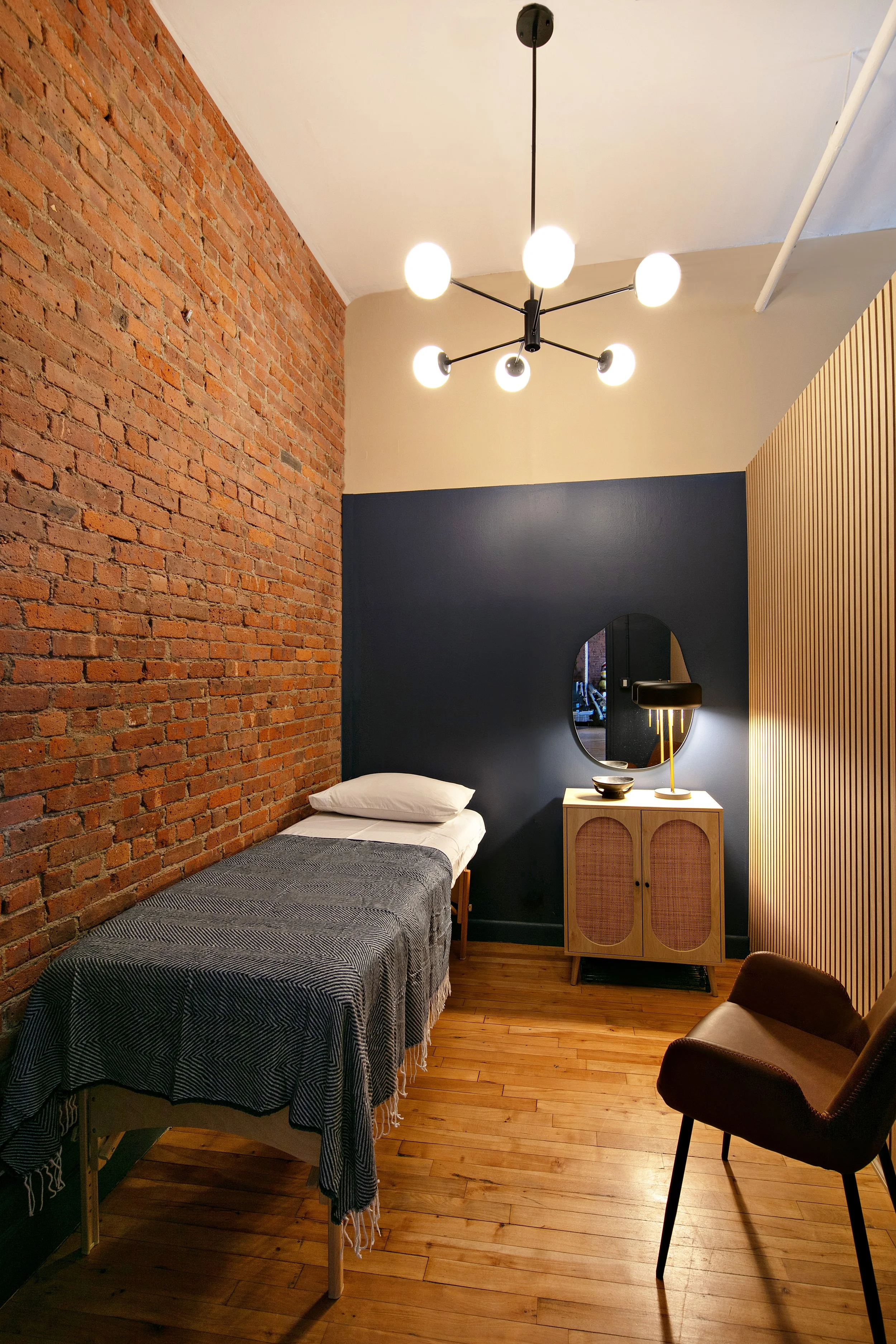 Minimalist bedroom with single bed, exposed brick wall, modern chandelier, wooden floor, small cabinet with mirror and lamp, and a chair.