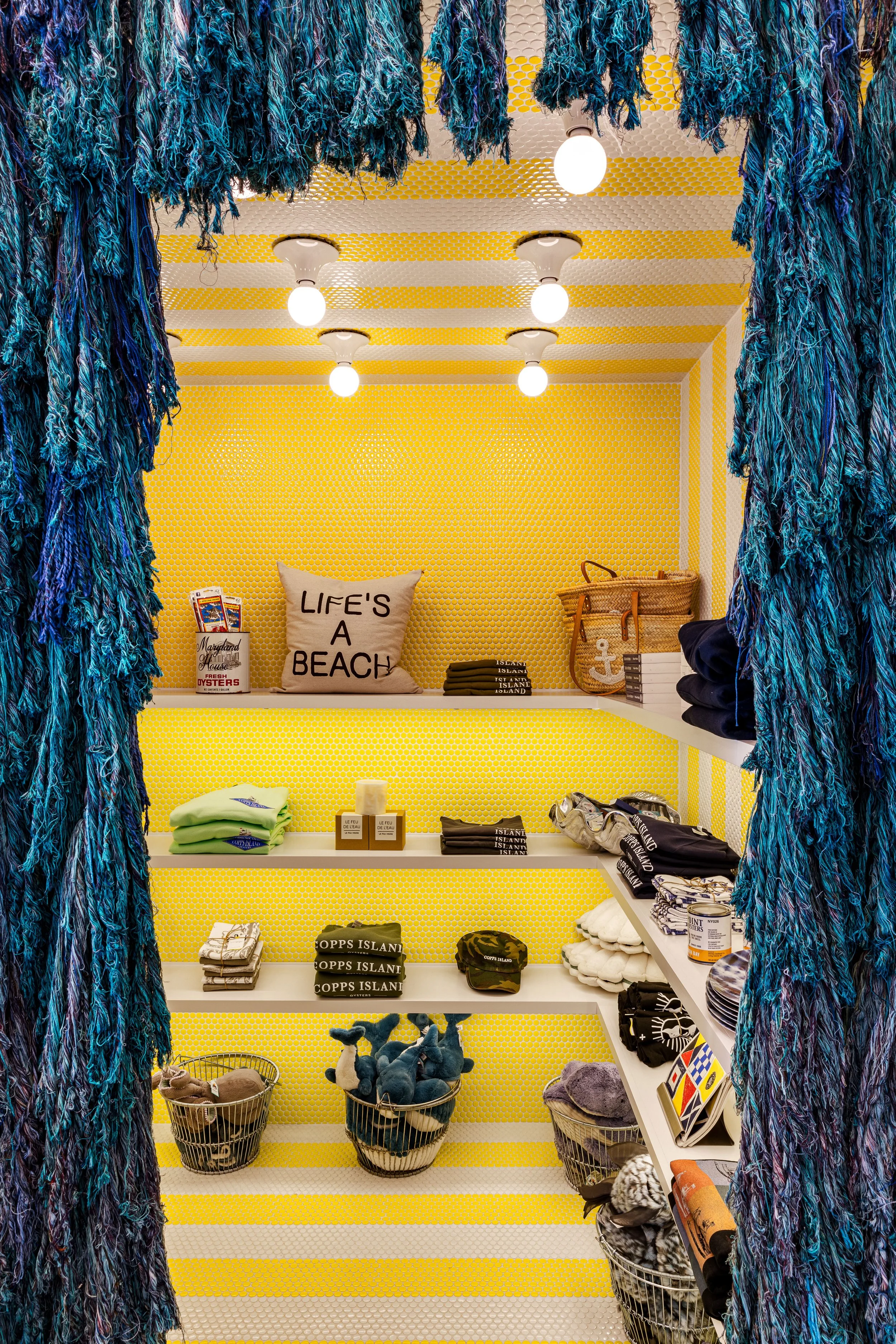 A boutique display with yellow walls featuring beach-themed merchandise, including shirts, pillows, baskets with stuffed animals, and accessories. Blue and green fabric decor hangs from the top.