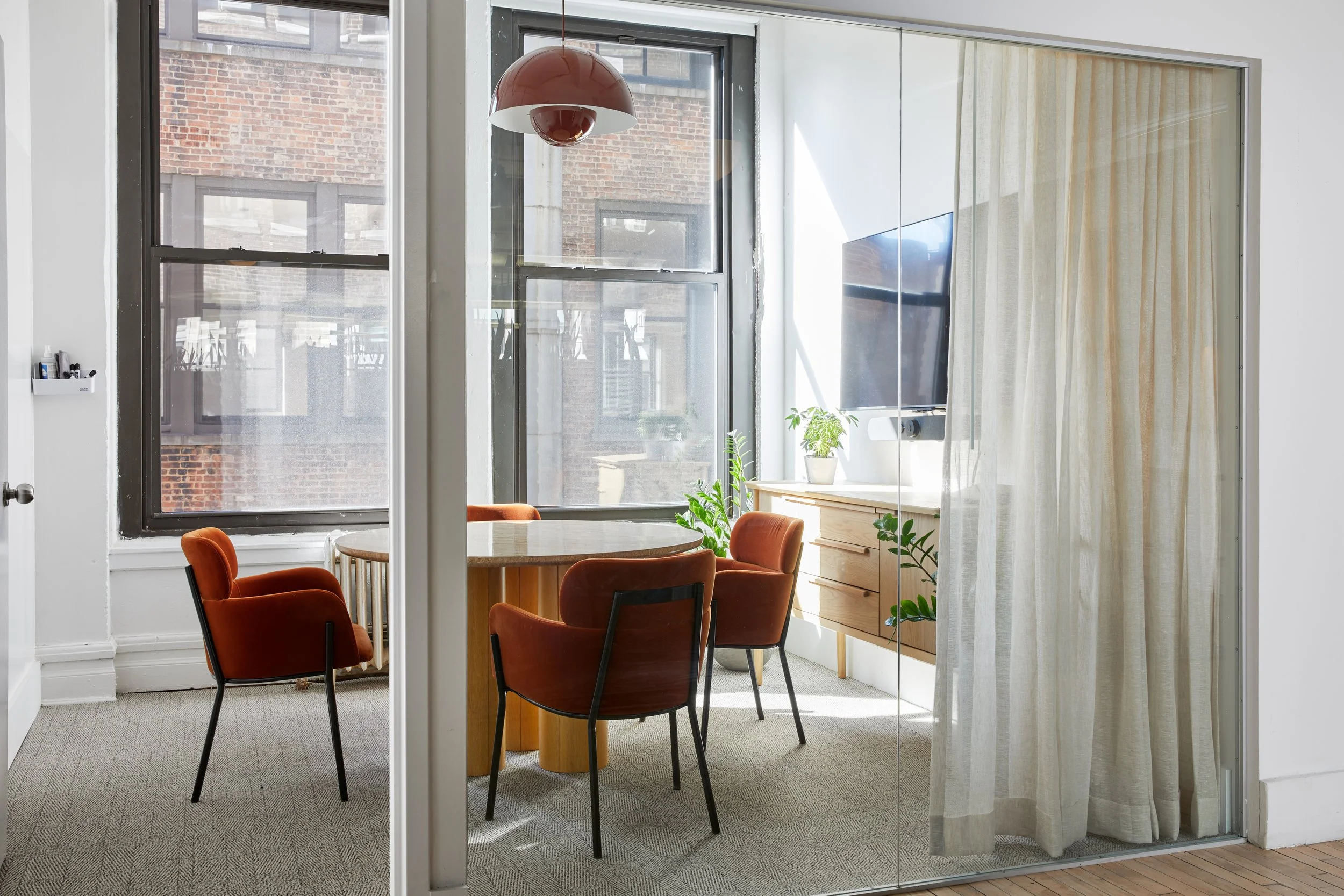 Modern conference room with round table, orange chairs, large windows, potted plants, television screen, and wooden sideboard.