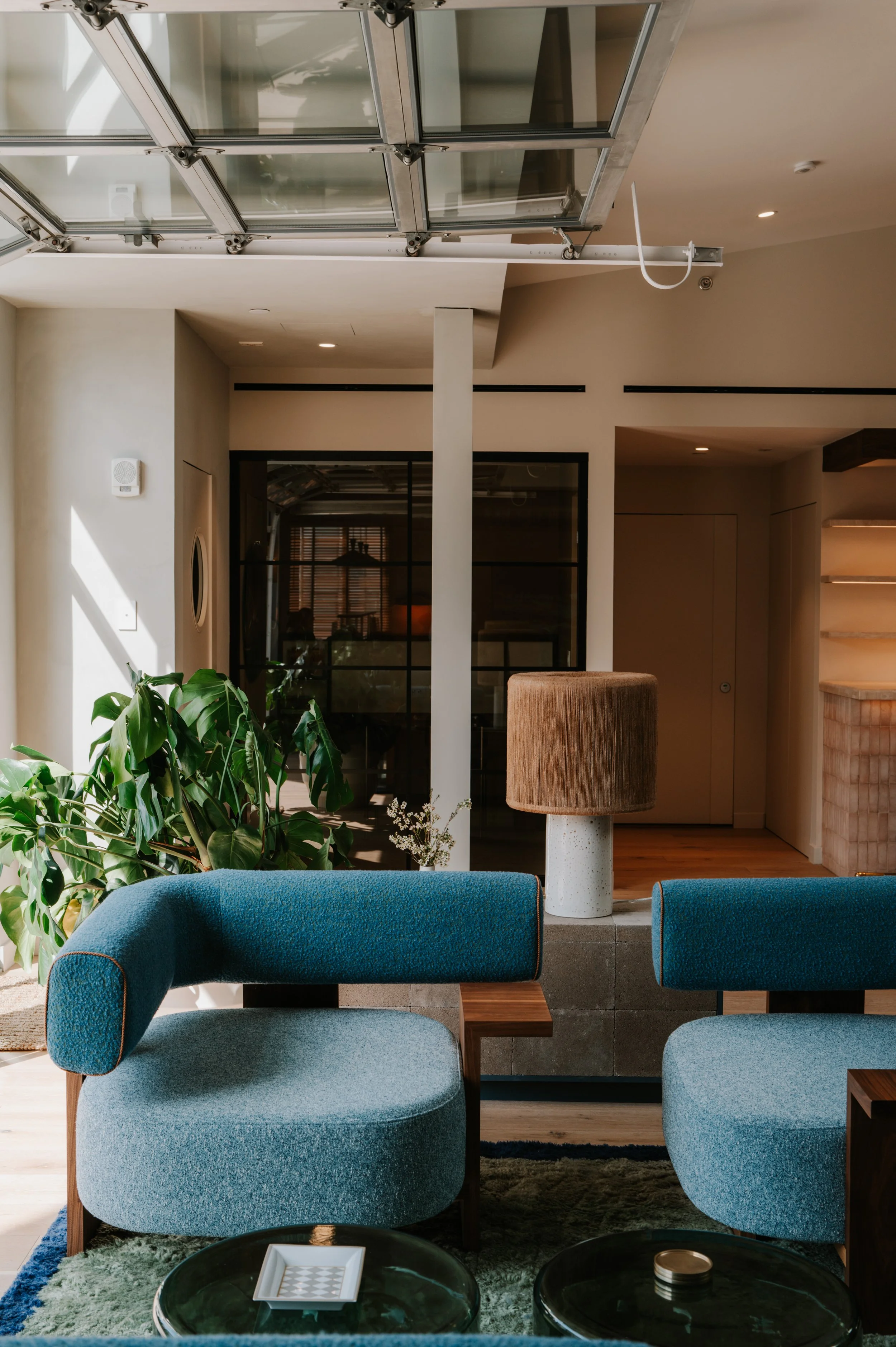 Modern living room with blue upholstered chairs, green plant, brown lampshade on white base, glass coffee table, and a balcony door reflected in glass wall.