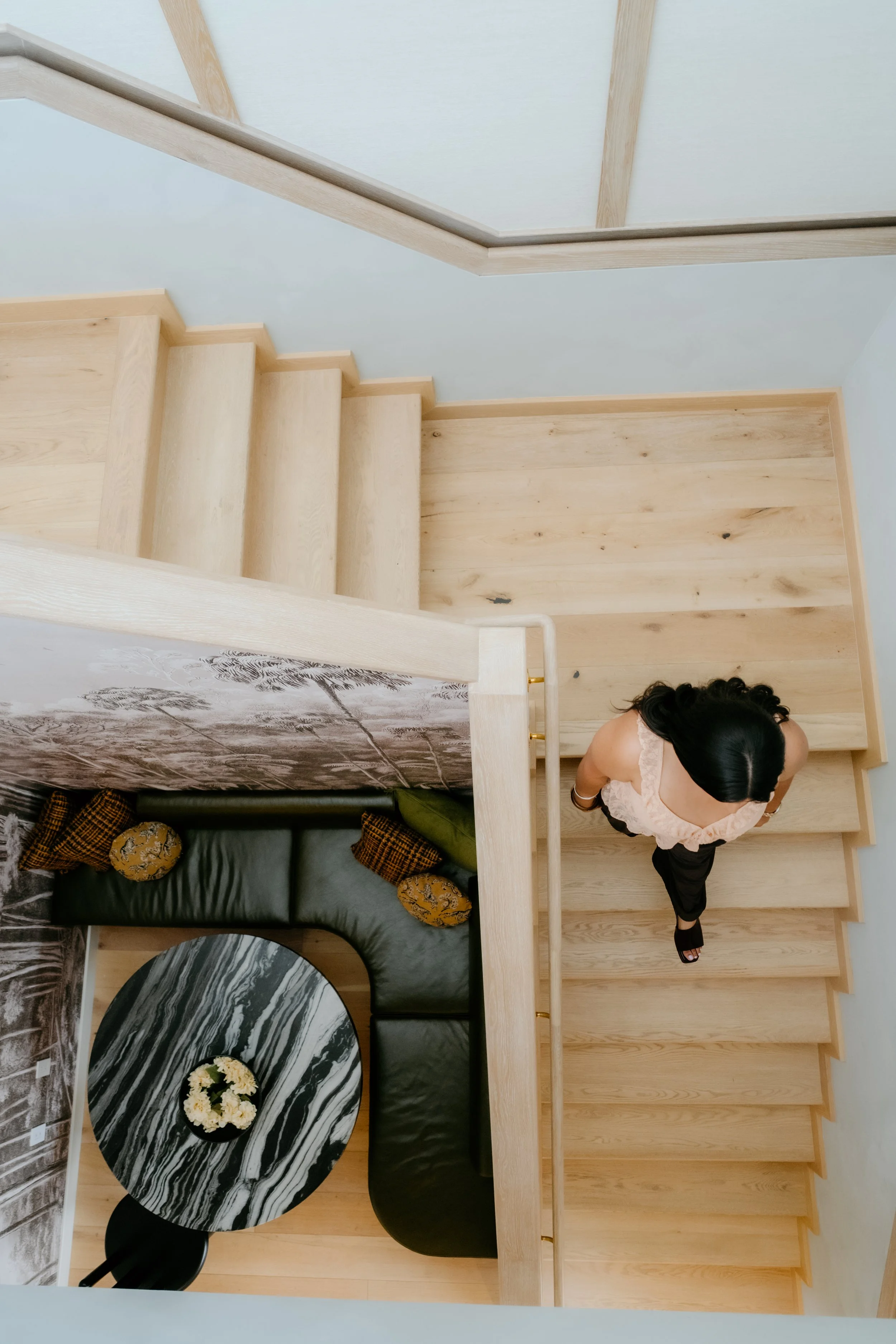 Overhead view of a woman walking down wooden stairs in a modern home, with a sitting area below featuring a black sofa and a round marble table.
