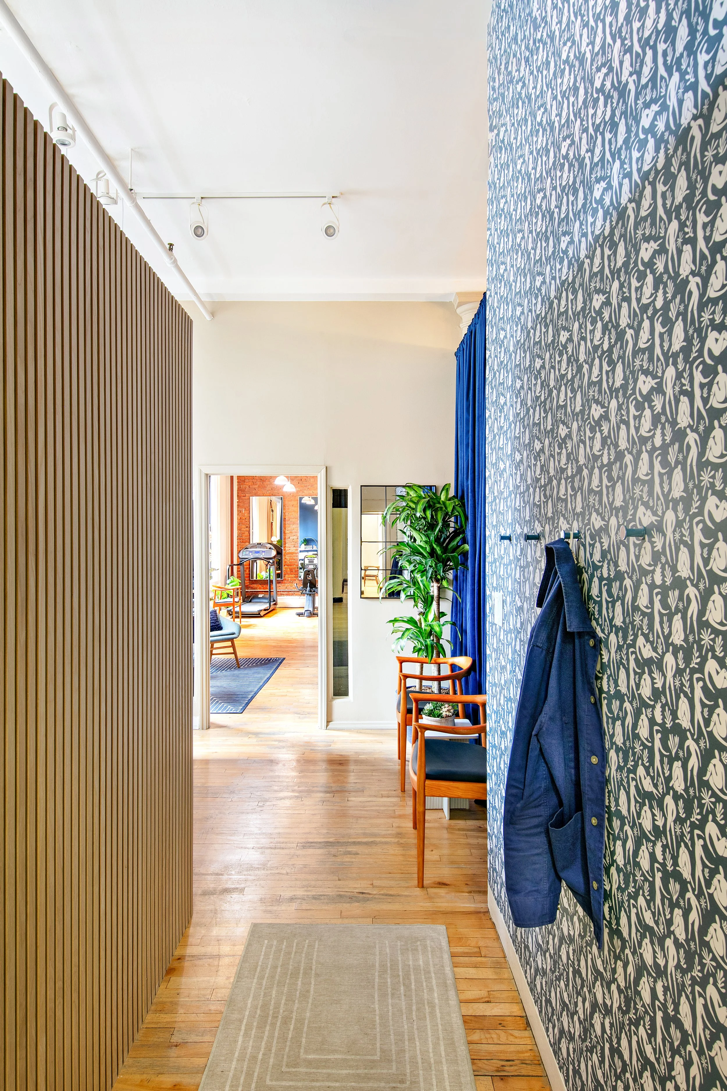 Well-decorated hallway with a patterned wall, wooden floor, rug, hanging coat, chairs, and a plant; leading to a room with exercise equipment.