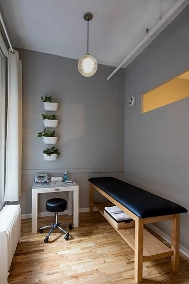 Small doctor's office with an examination table, white desk, black stool, and wall-mounted white planters.