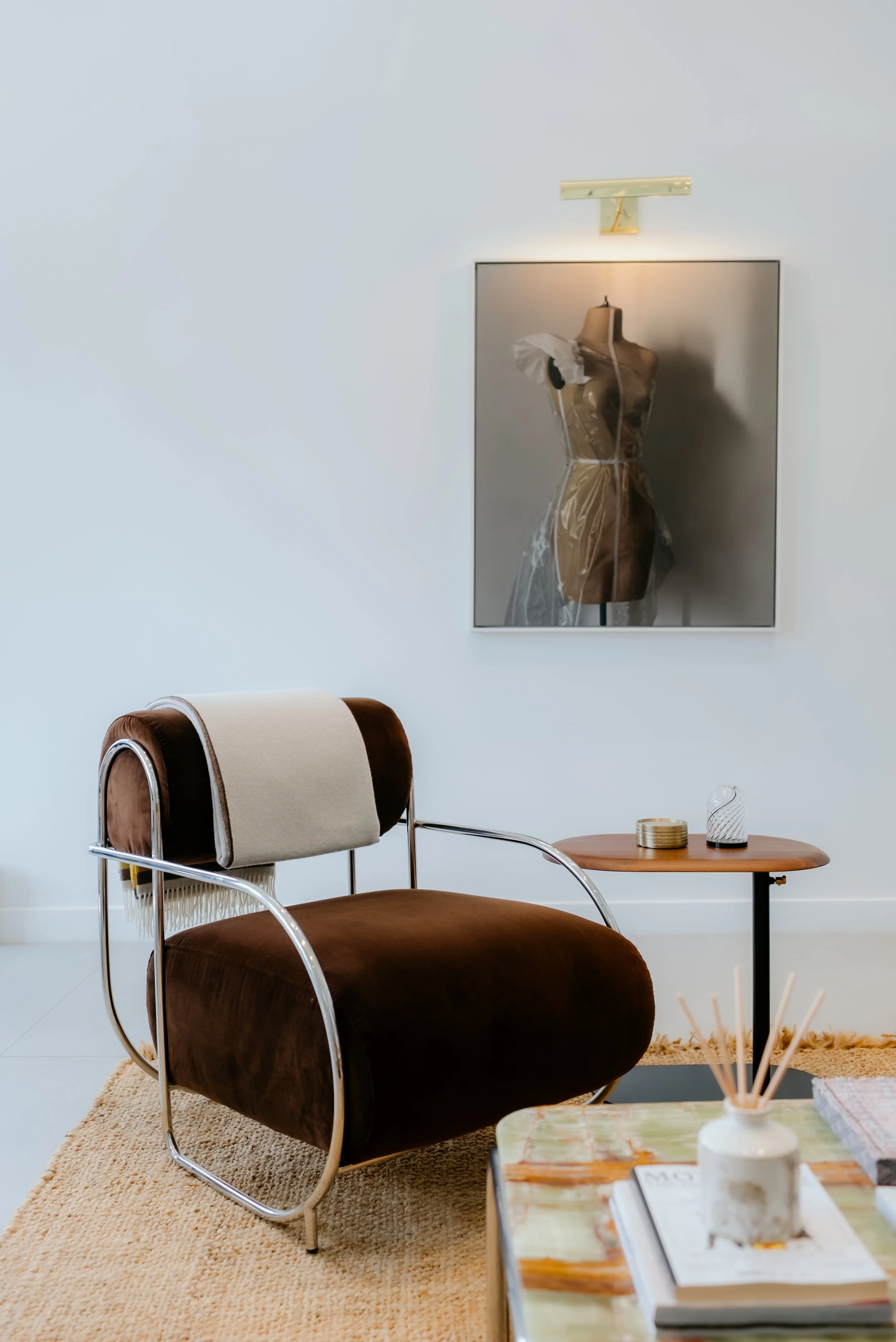 Modern interior with a brown velvet armchair, a beige throw blanket, a wooden side table with a coaster and decorative glass, a white vase with reed sticks, and a framed photo of a dress form on the wall.