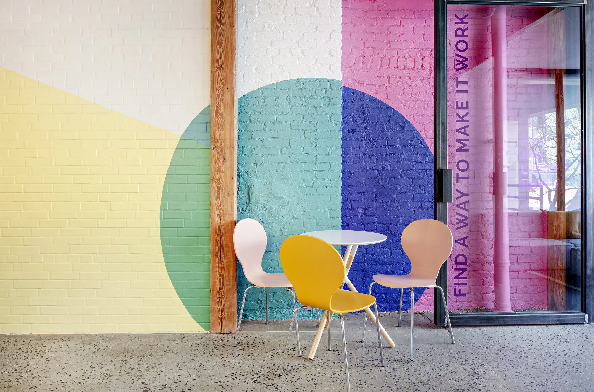 Colorful office space with abstract painted walls, featuring pastel shades of yellow, green, blue, and pink. The room includes a small round table with four modern chairs in pastel colors. A glass door has the phrase 'Find a way to make it work' written on it.