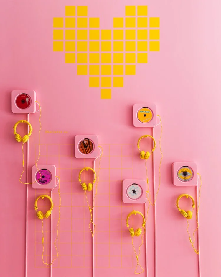 Pink wall with yellow heart made of squares, featuring yellow headphones and CDs in pink holders.