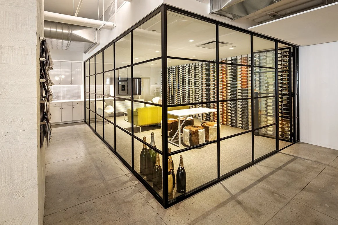 Modern glass-enclosed office space with wine display, table, seating, and industrial-style ceiling.
