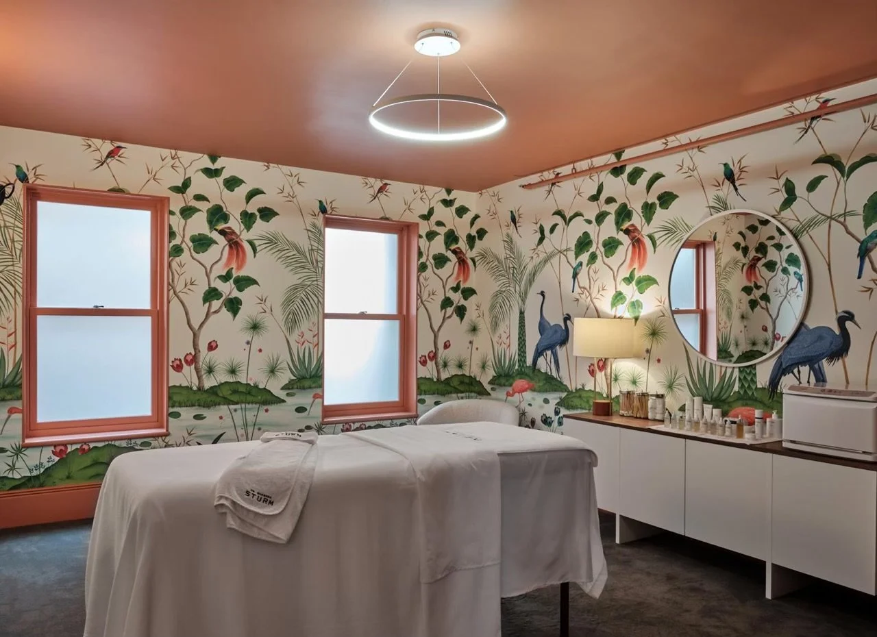 Spa treatment room with floral wallpaper, massage table covered in white sheets, a circular ceiling light, two windows, a lamp, a mirror, and skincare products on a counter.