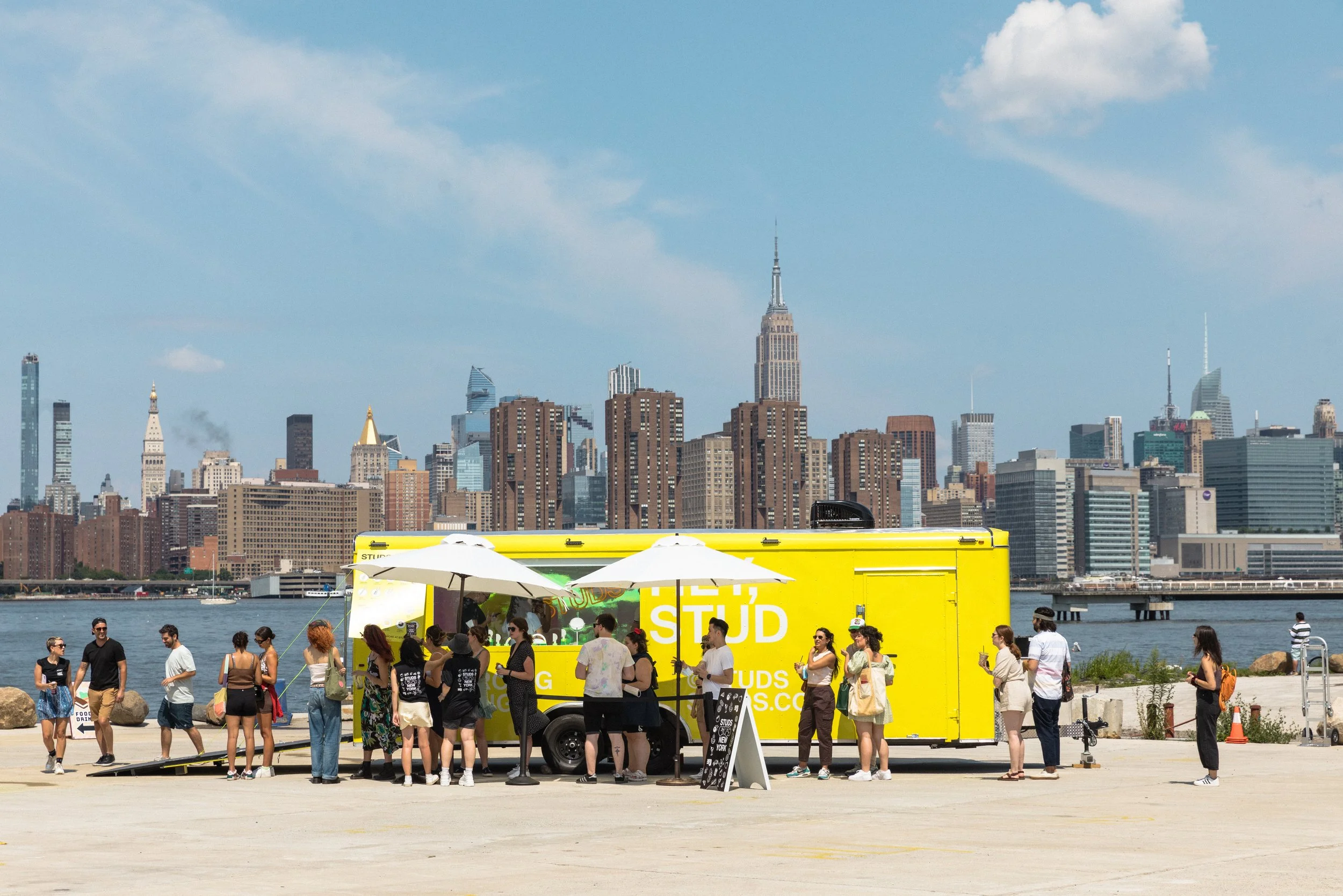 People gathering near a bright yellow food truck on a waterfront with a city skyline in the background under a blue sky.
