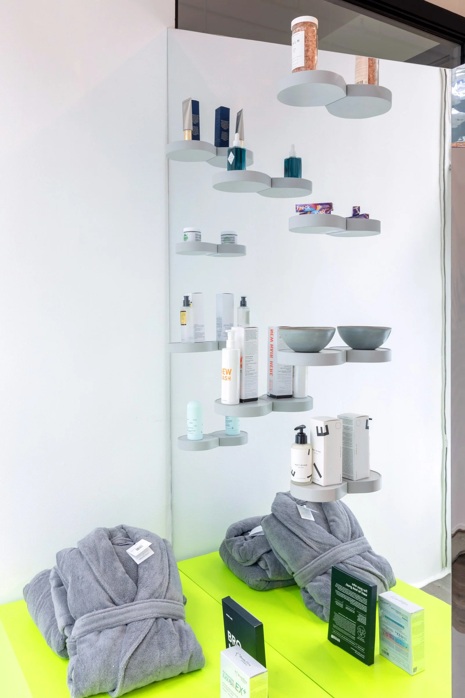 Display of skincare and wellness products on floating shelves, including bottles, creams, and boxes, with gray towels on a neon yellow table.