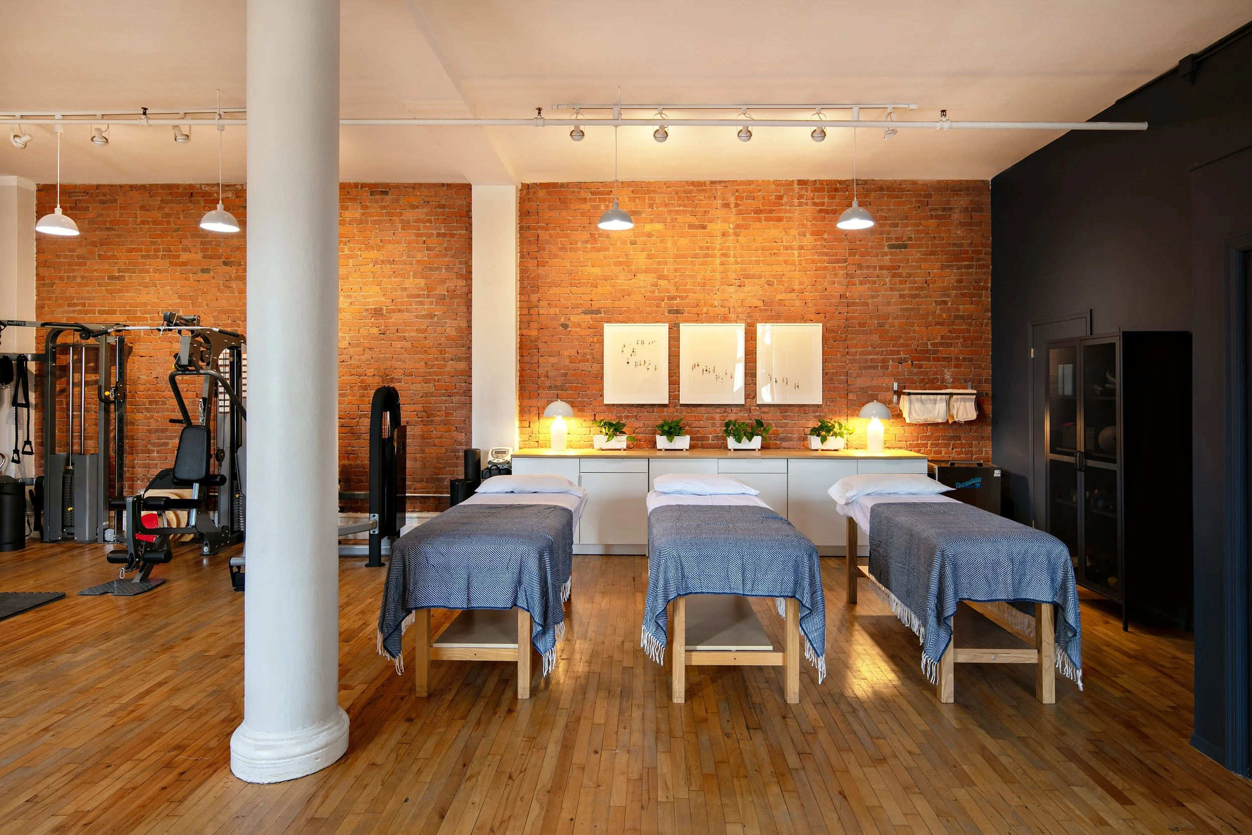 Interior of a spa with three massage tables covered in gray blankets, a brick wall with framed art, potted plants, and ambient lighting. Gym equipment is visible in the background.