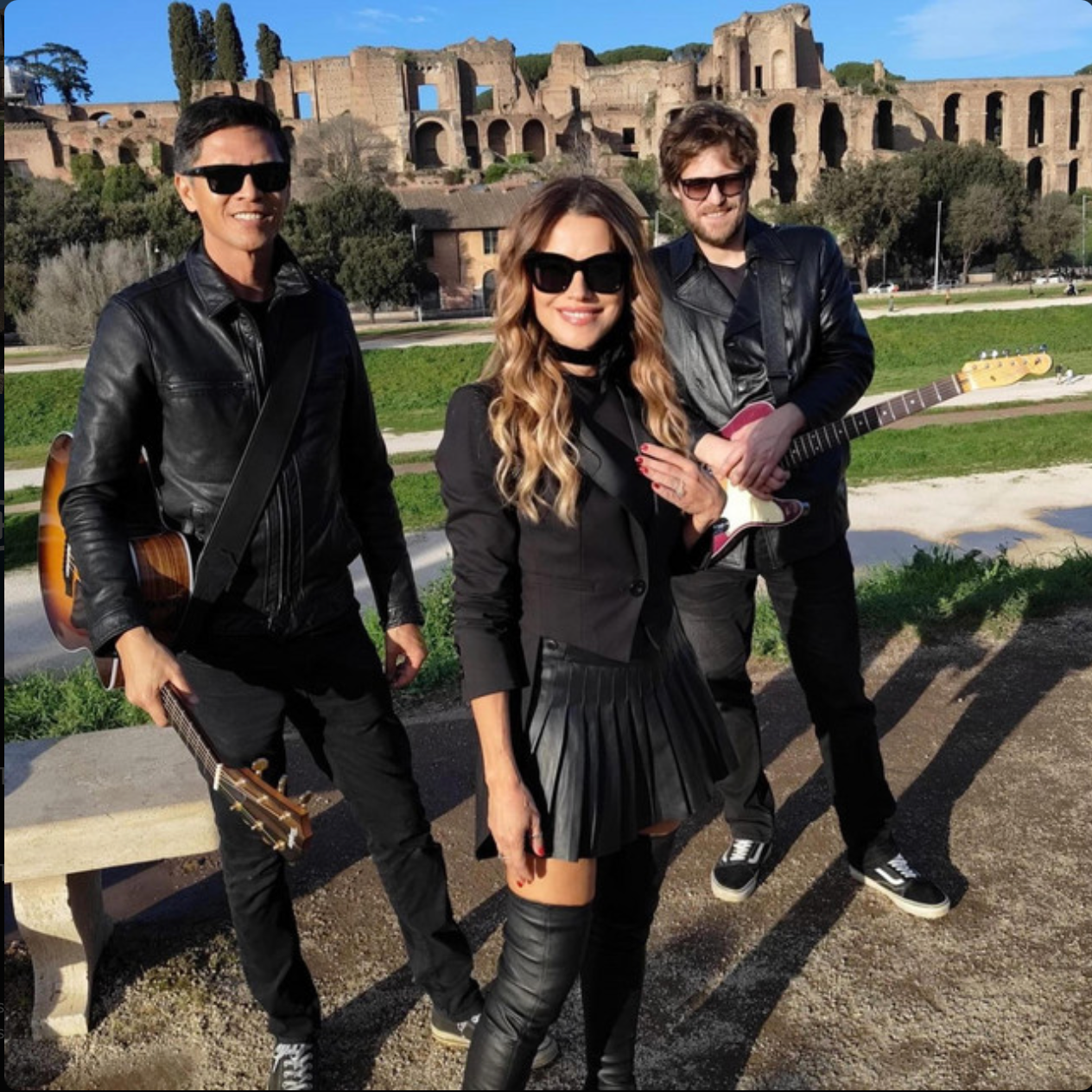 Three musicians with guitars in front of the Roman ruins of the Colosseum, wearing sunglasses and black leather jackets, standing on a pathway with green grass and trees.