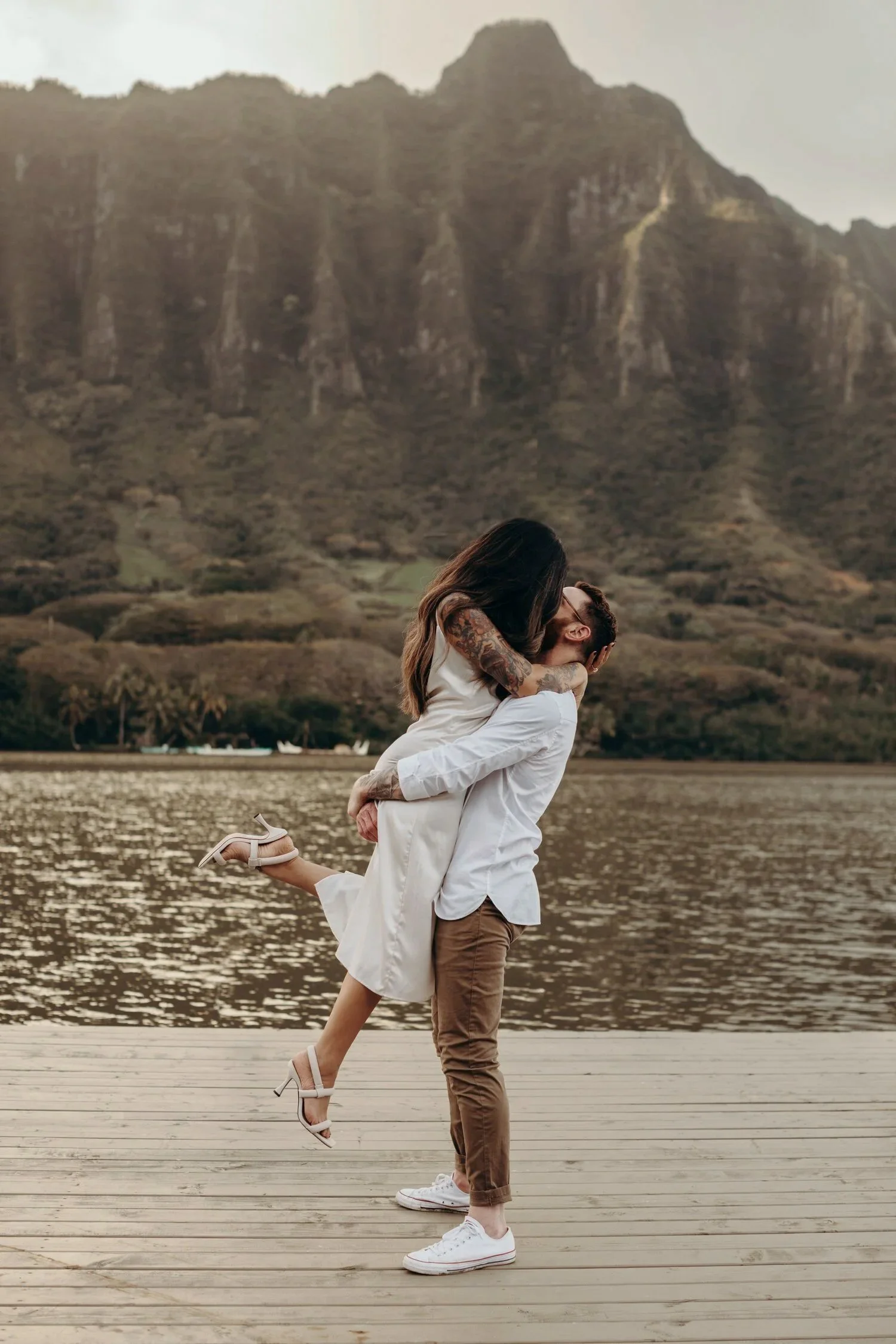 An engagement session at Kualoa Ranch. The woman is being lifted and kissed by the man, wearing white and beige clothing.