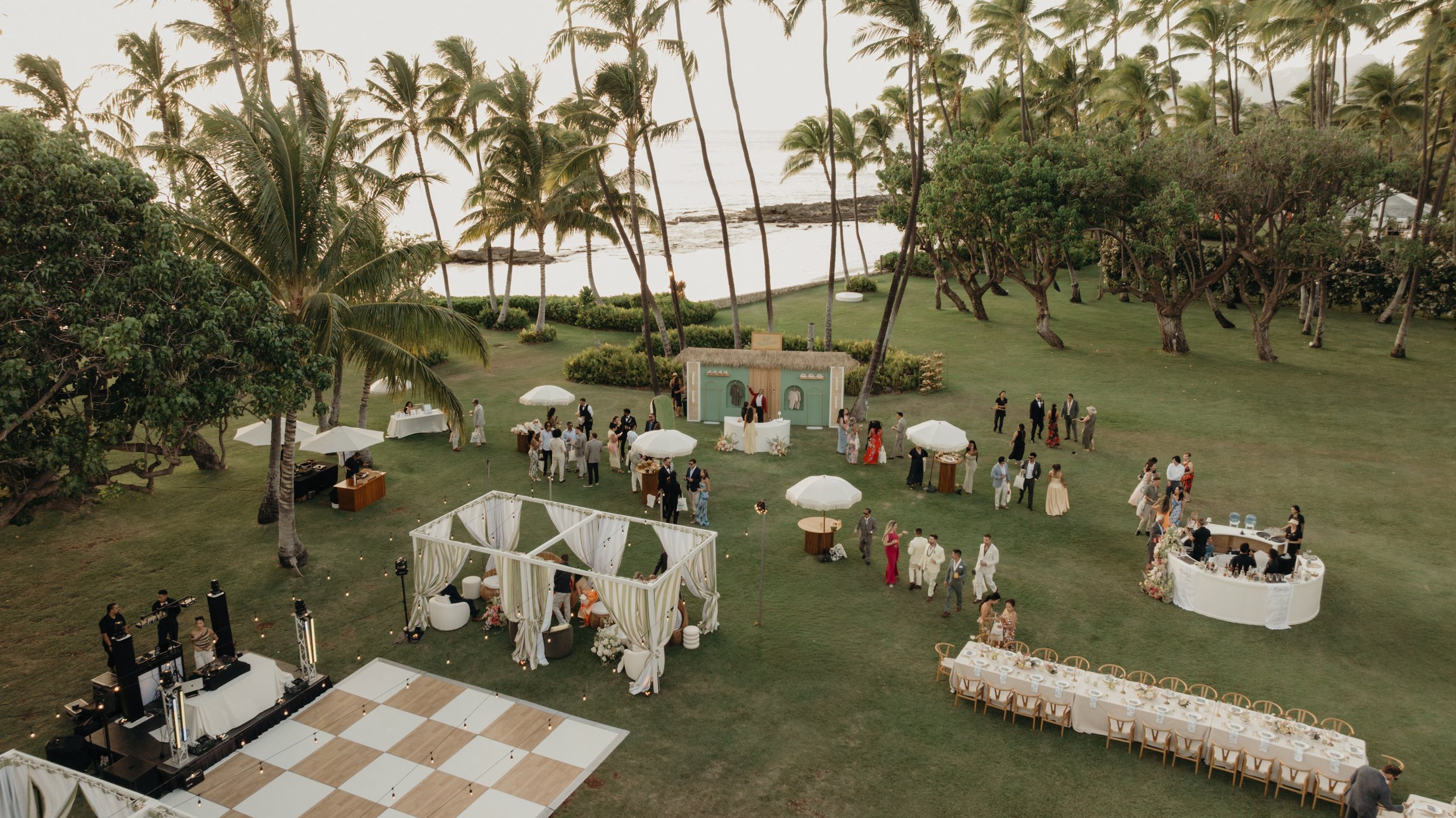 Hawaii destination wedding reception at Lanikuhonua. Outdoor wedding on a grassy lawn near the beach with palm trees, tables, umbrellas, a dance floor, and decorated stage, with guests socializing and a band performing.