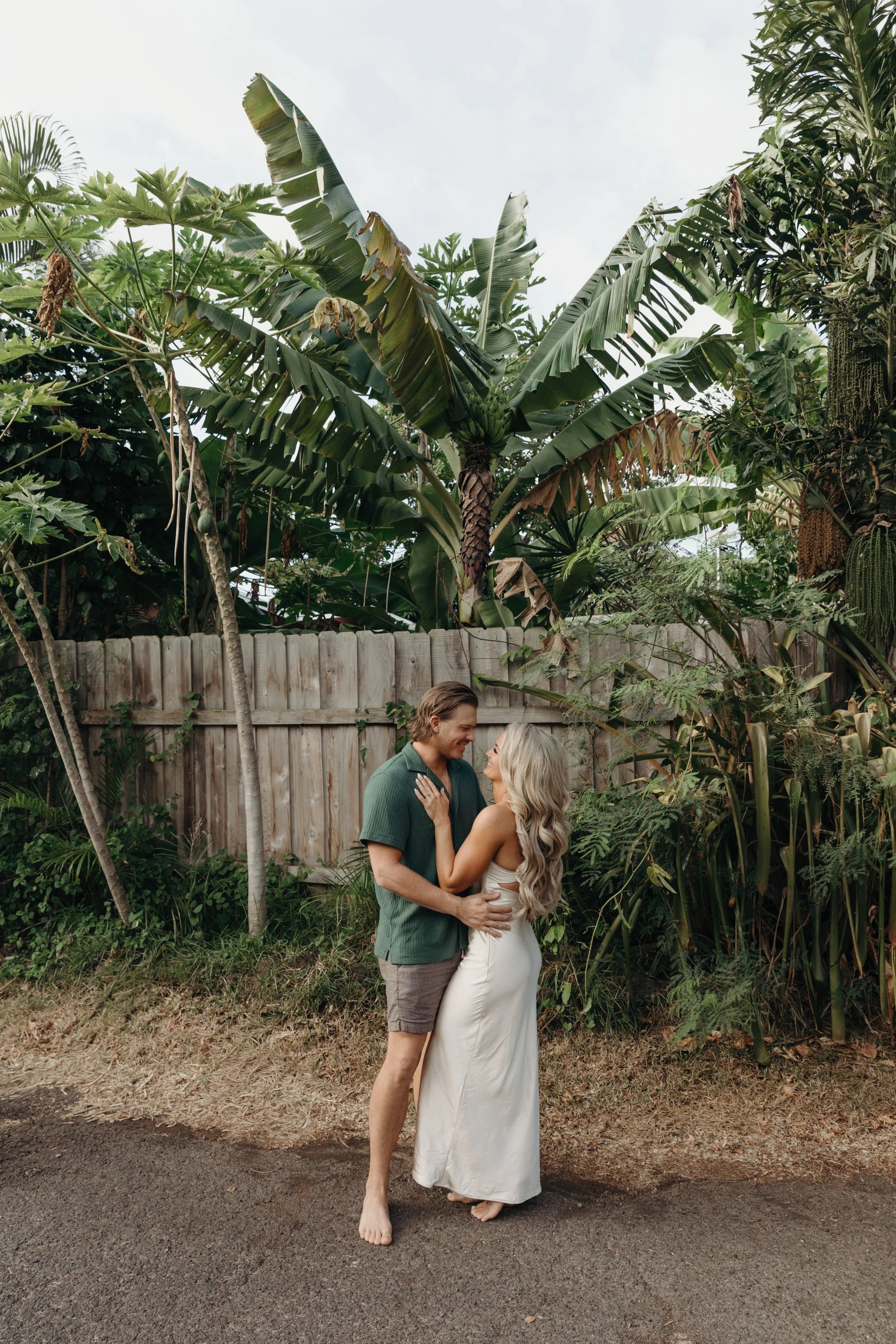 A couple stands close together in a garden, holding hands and looking at each other happily. The woman has long, curly blonde hair and is wearing a white dress, while the man has short brown hair and is wearing a green shirt and shorts. They are bare