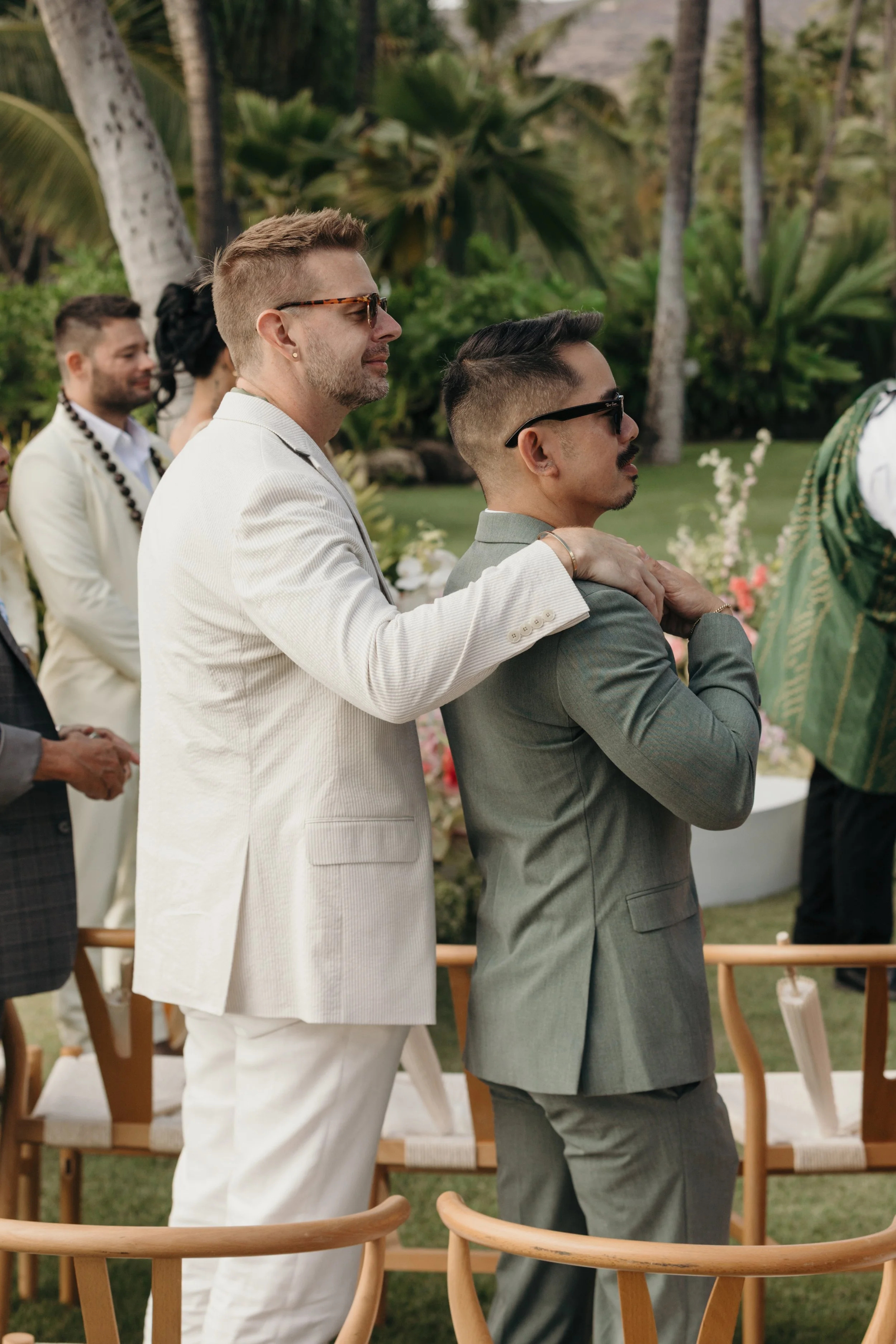 Two men in suits, one with a white suit and another with a gray suit, standing close together during an outdoor wedding ceremony with tropical trees in the background.