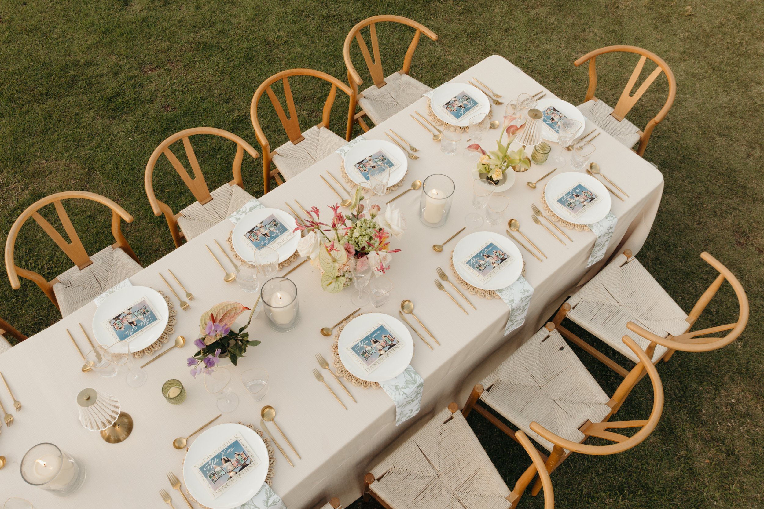 An outdoor rectangular dining table set for eight, with a white tablecloth, gold flatware, clear glasses, and white plates with a colorful design, accompanied by wooden chairs with woven seats. The table is decorated with flowers, candles, and small 