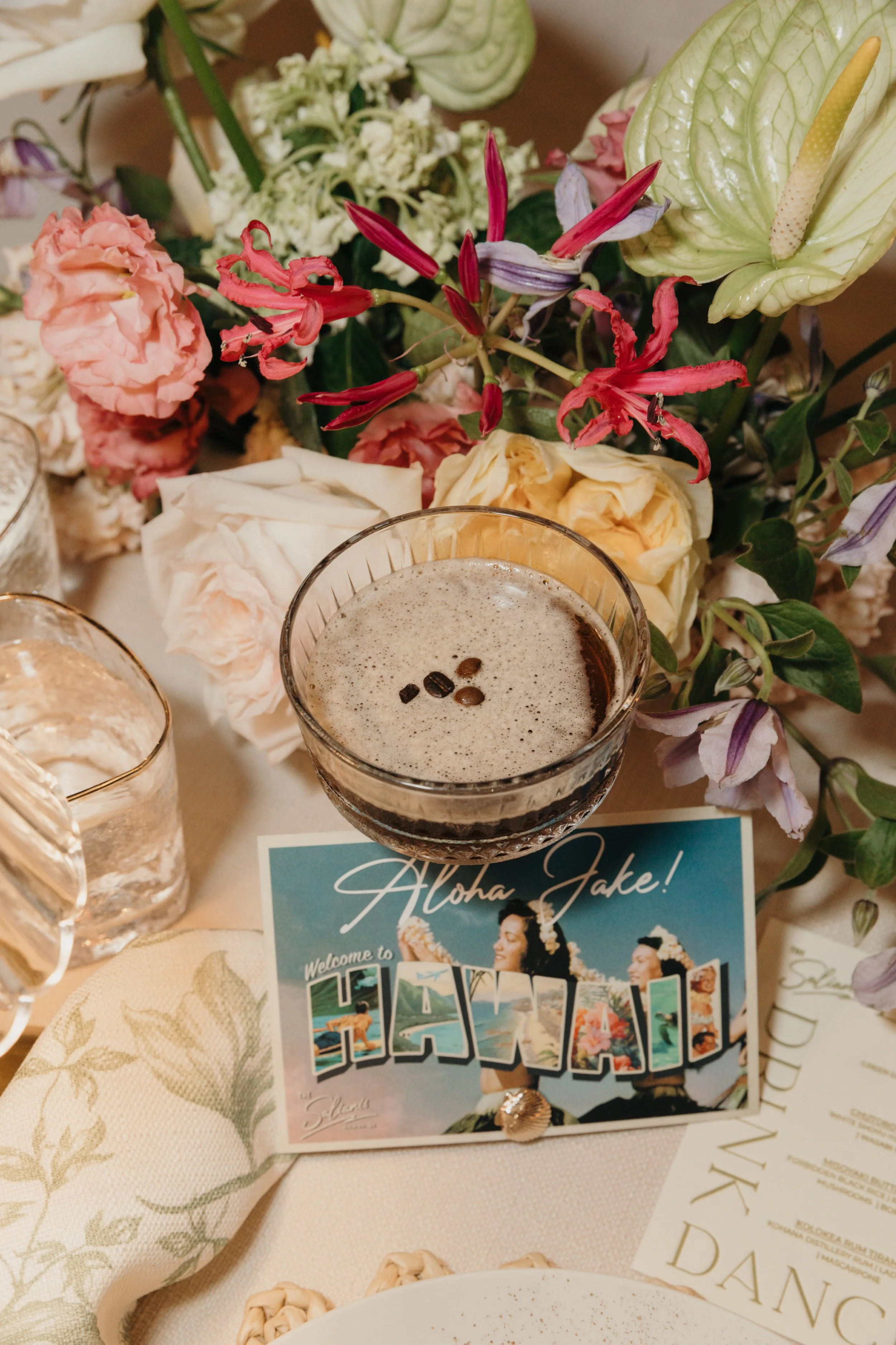 An espresso martini surrounded by colorful flowers, a Hawaiian-themed postcard, and decorative items on a table.