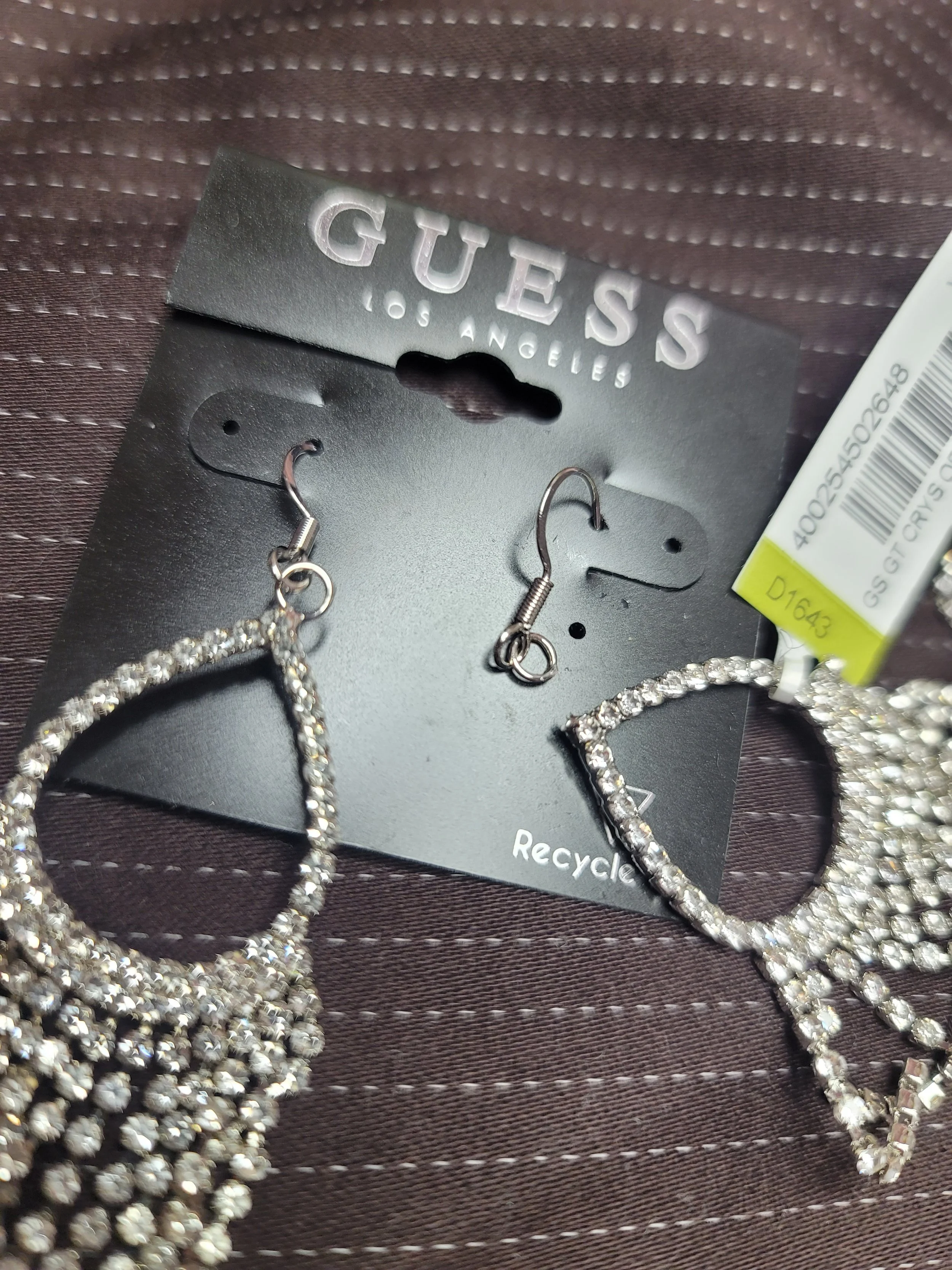 A pair of Guess jewelry earrings on a black display card with rhinestones, placed on a brown textured surface.