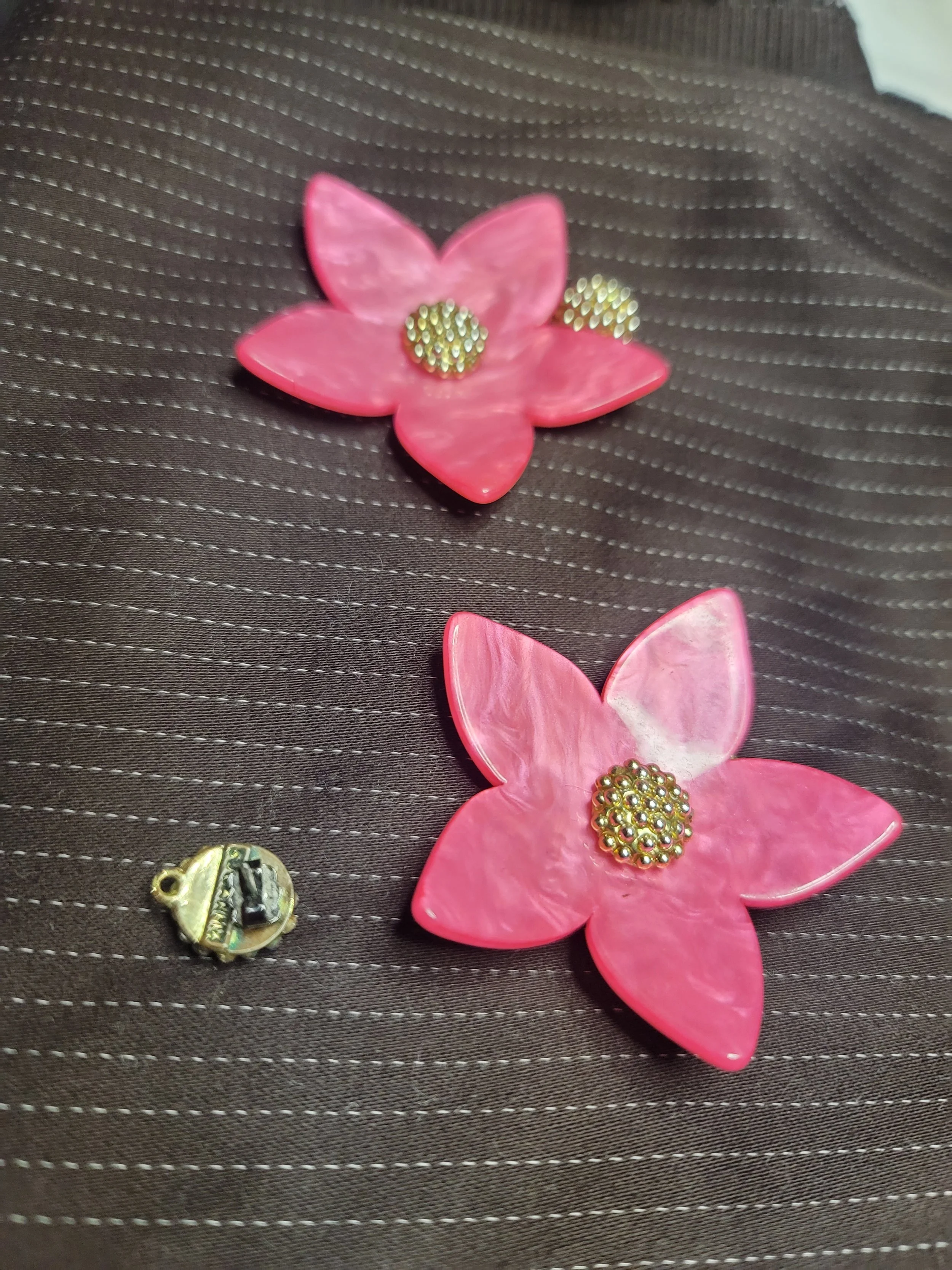 Two pink flower-shaped brooches with pearl-studded centers and a small metallic clasp, placed on a brown fabric surface with white thread pattern.
