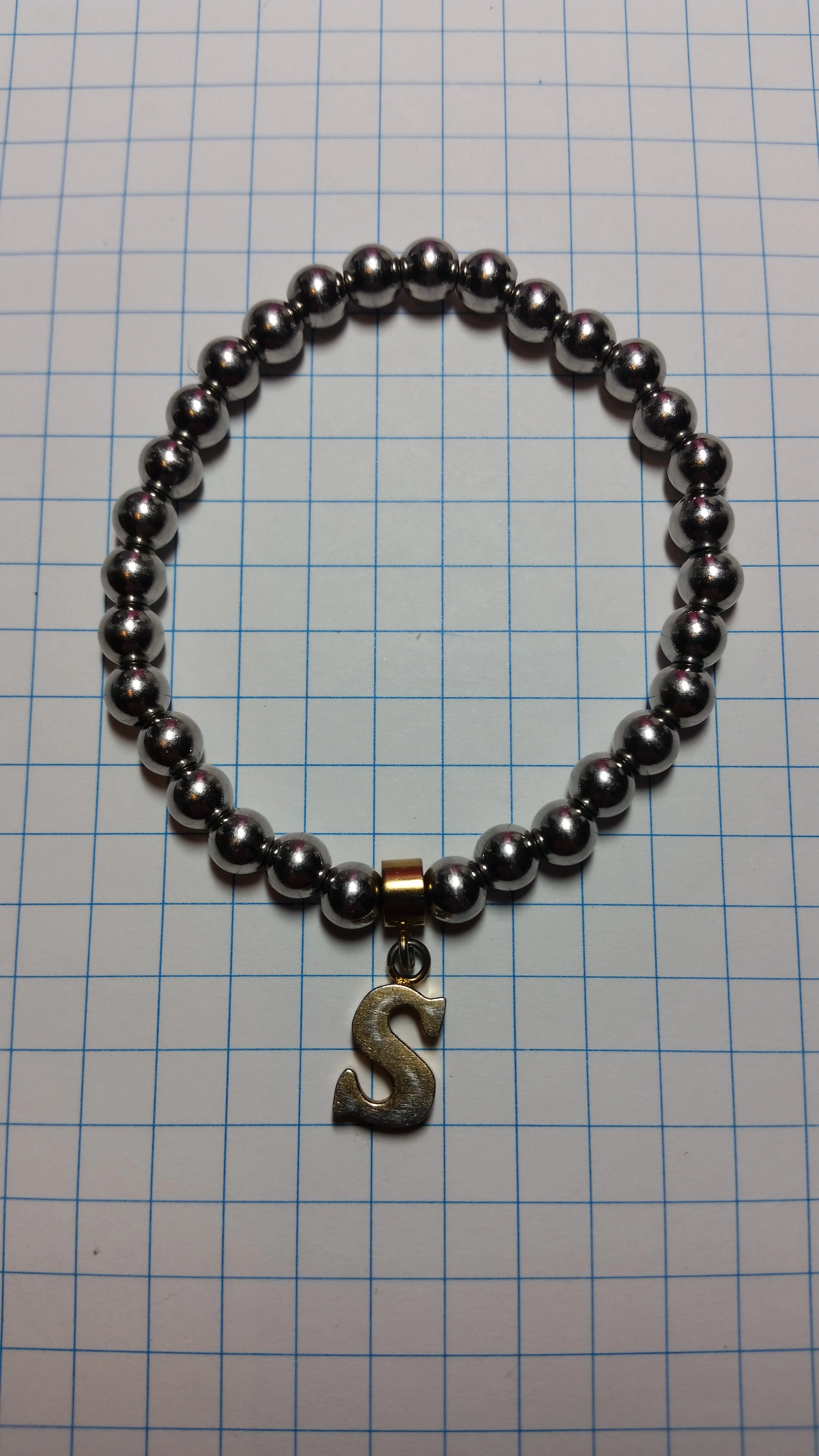 A beaded bracelet with metallic silver beads and a gold letter 'S' charm, placed on a blue graph paper background.