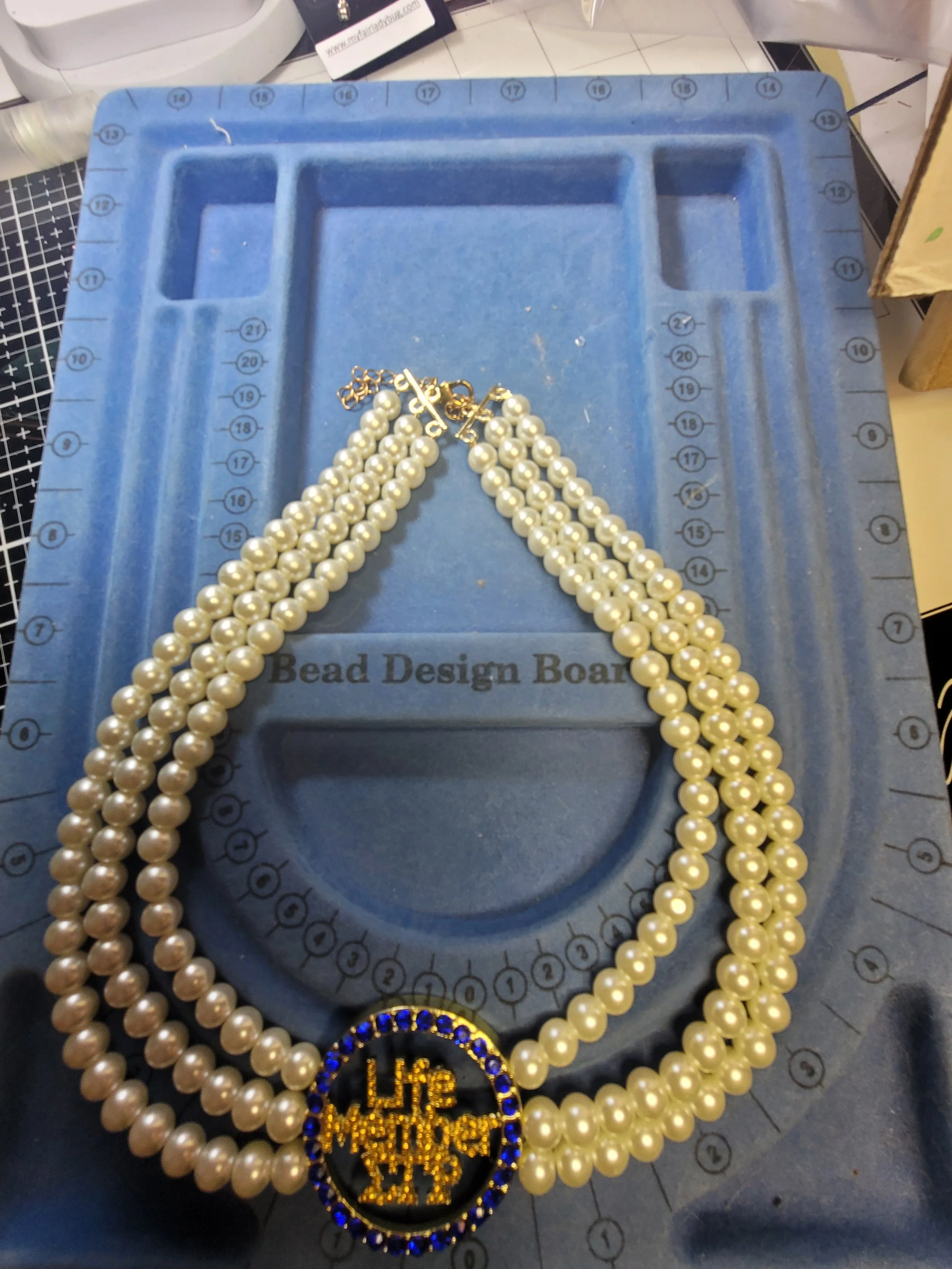 A pearl necklace with a circular gold and blue 'LIFE MESSAGE' pendant on a blue bead design board.