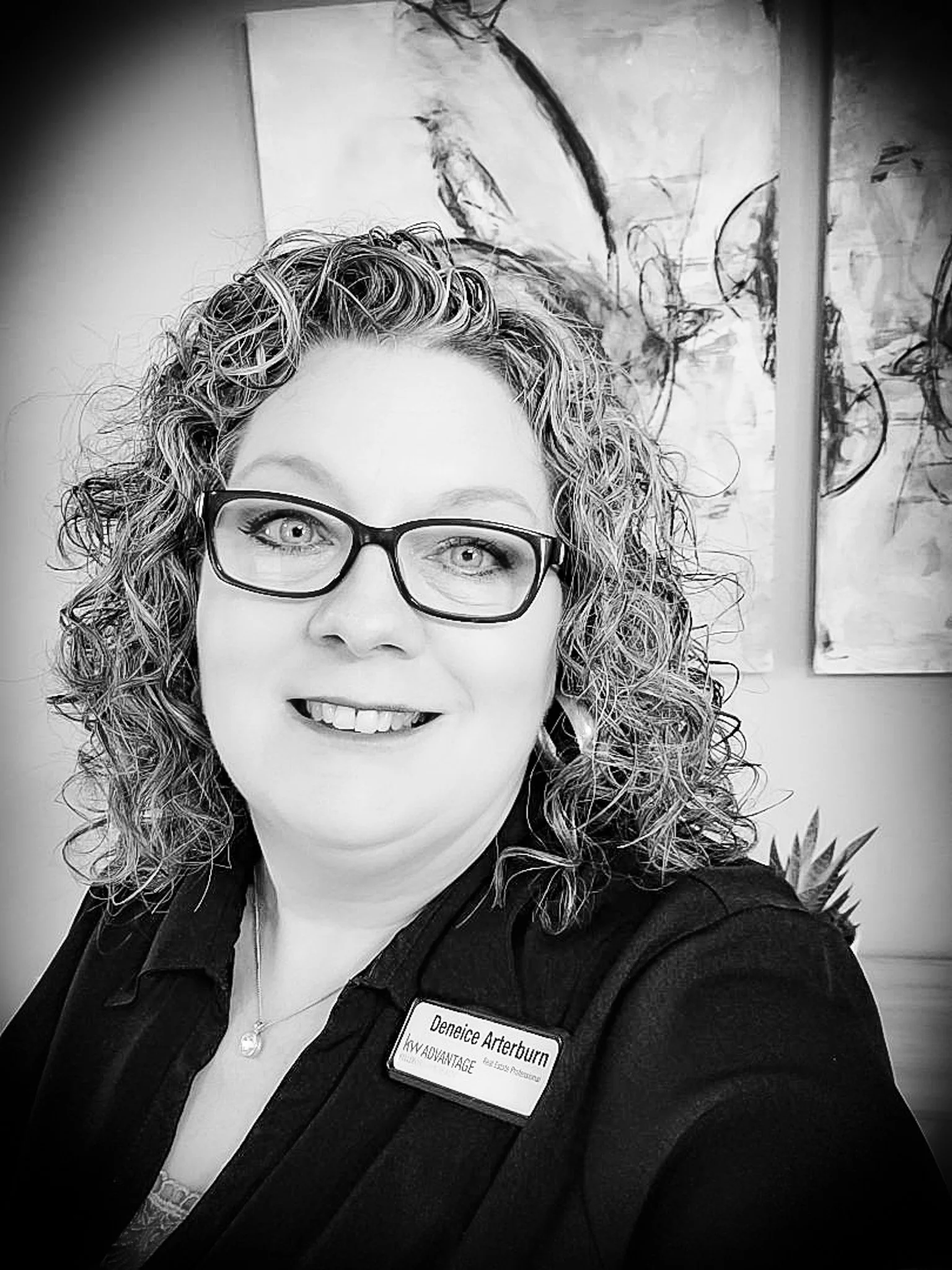 A woman with curly hair, glasses, and a friendly smile taking a selfie. She is wearing a name tag that reads "Deneice Arterburn, KW Advantage, Realty, and Associates." Background includes abstract artwork and a plant.