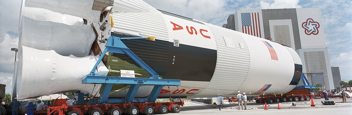 Saturn V Rocket Moving to the Saturn V Center, with the Vehicle Assembly Building in the Background