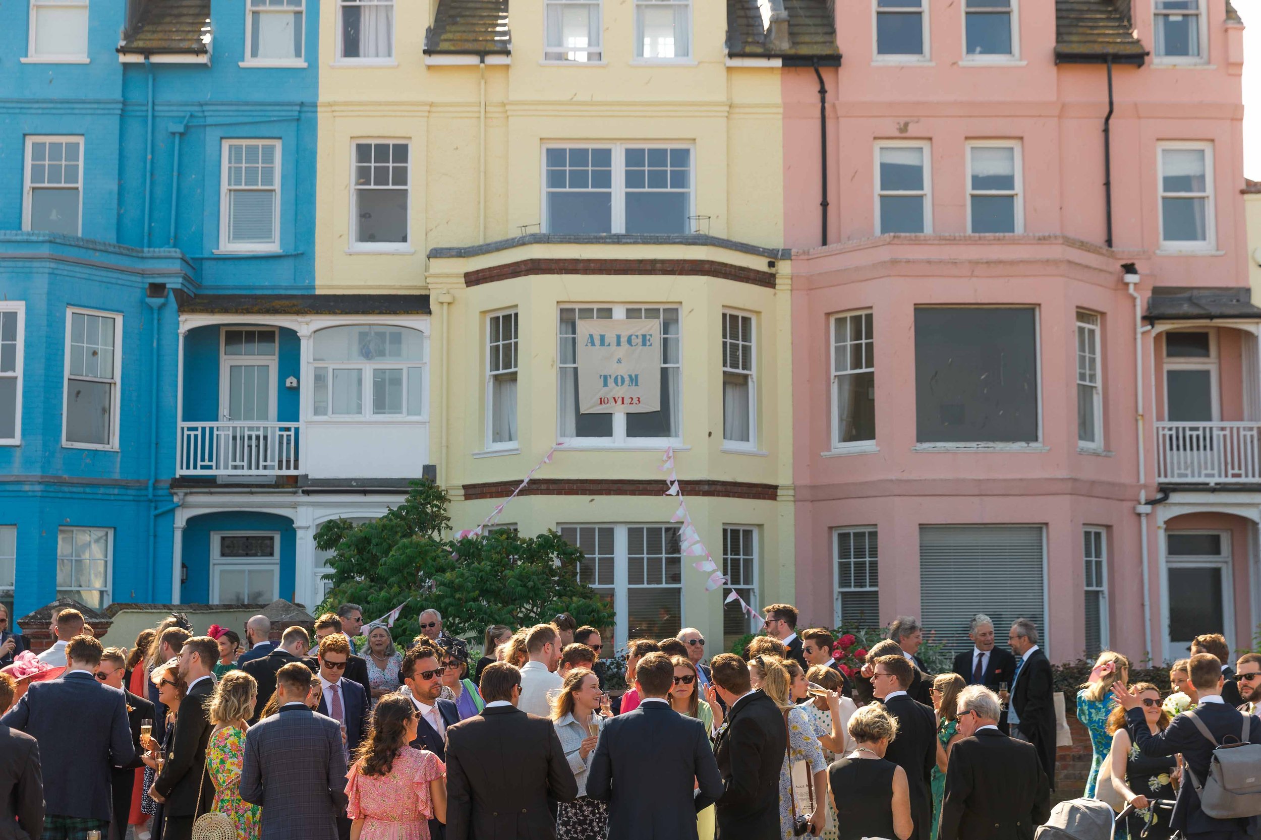 A crowd of people dressed in formal attire gathered outdoors in front of colorful apartment buildings with pastel blue, yellow, and pink facades. A sign in the yellow building reads "ALICE & TOM 10.VI.23."