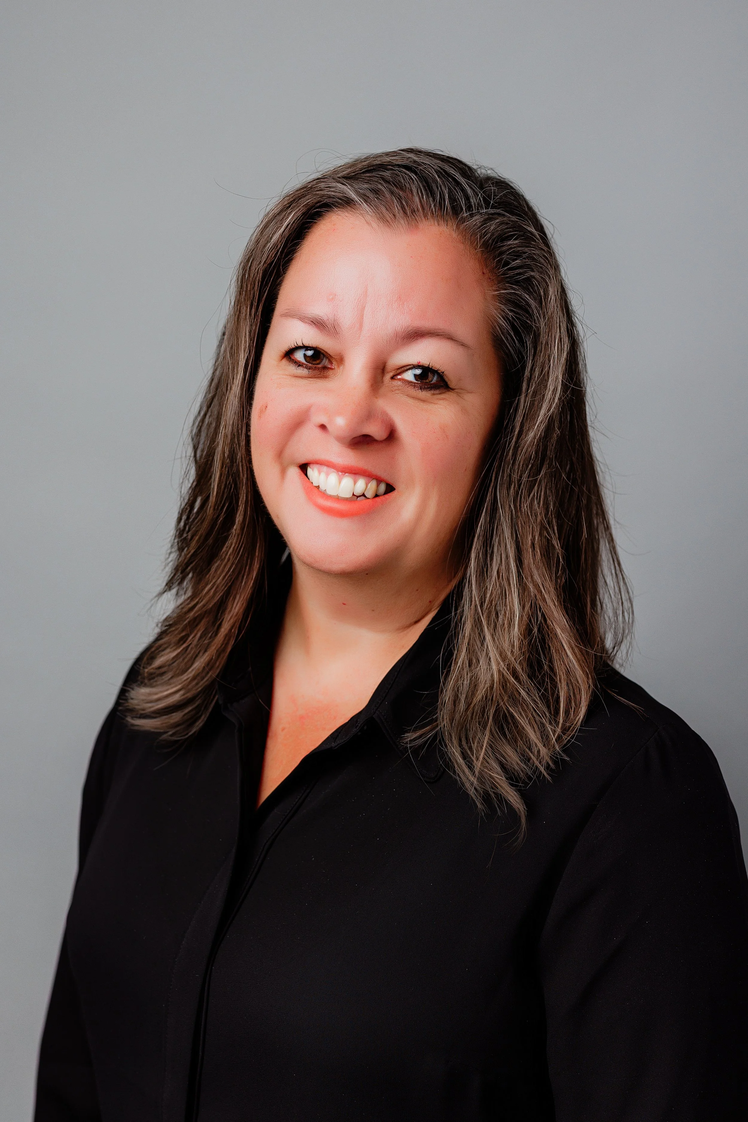 Smiling woman with shoulder-length hair wearing a black top, posing against a plain gray background.