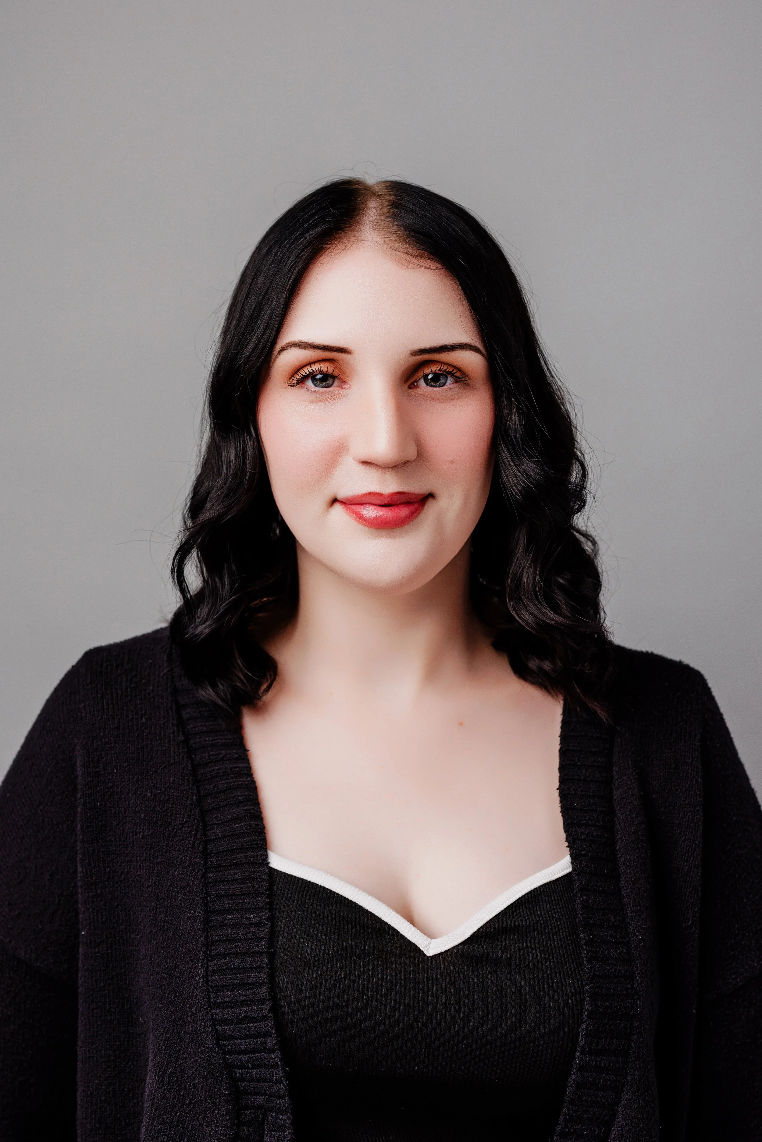 Portrait of a young woman with black shoulder-length wavy hair, fair skin, and red lipstick, wearing a black top with a white trim and a black cardigan, smiling softly against a plain gray background.