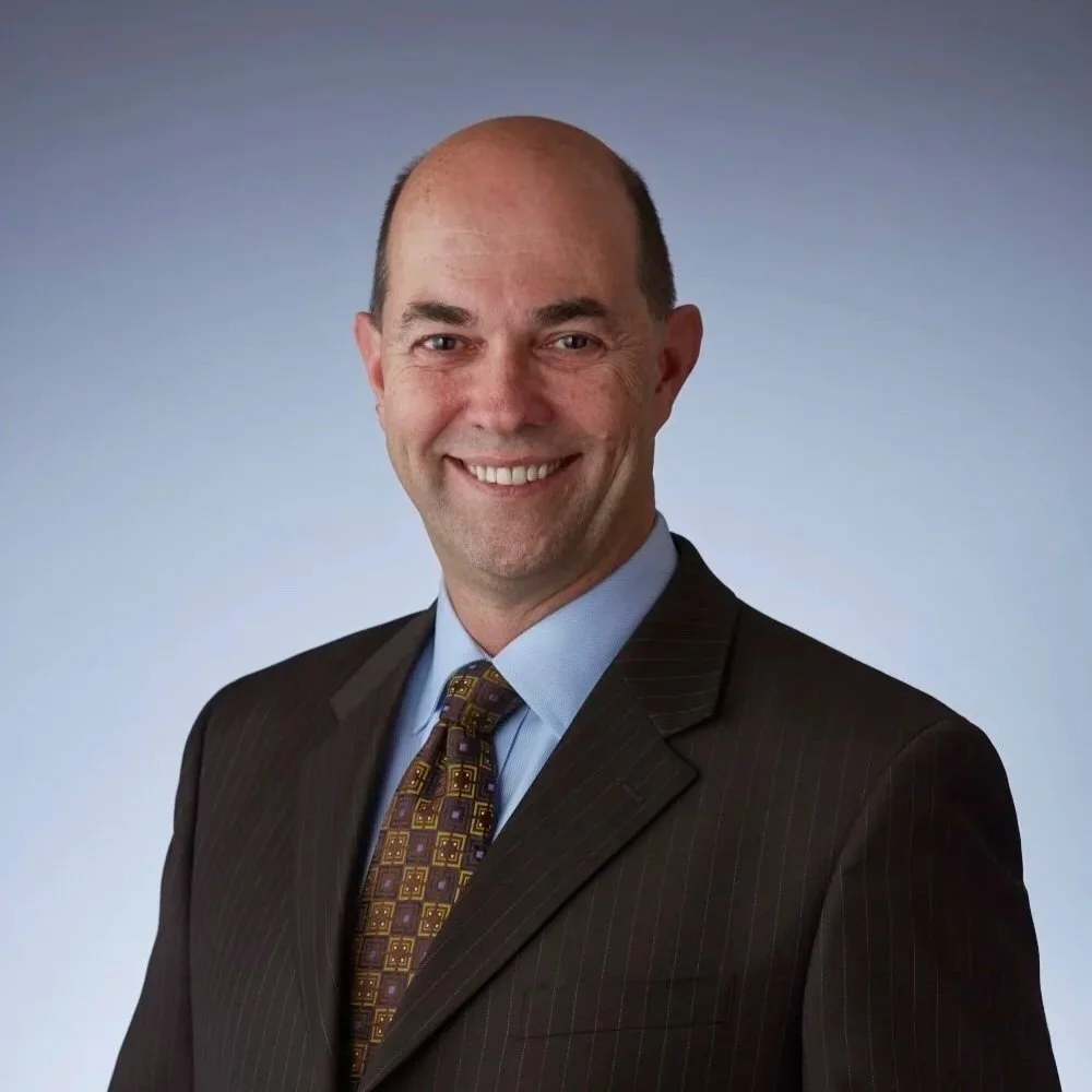 A smiling man in a dark pinstripe suit with a light blue shirt and patterned tie, against a plain gradient background.