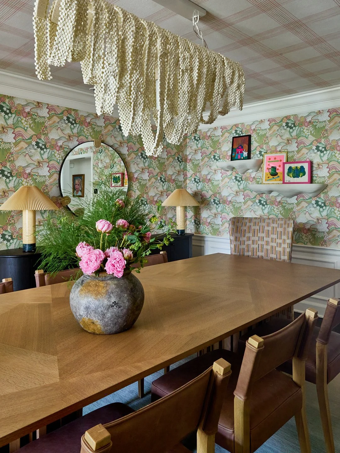 A dining room with a large wooden table, pink peonies in a rustic gray vase, two yellow lamps, decorative wallpaper, and colorful framed artwork on white shelves.