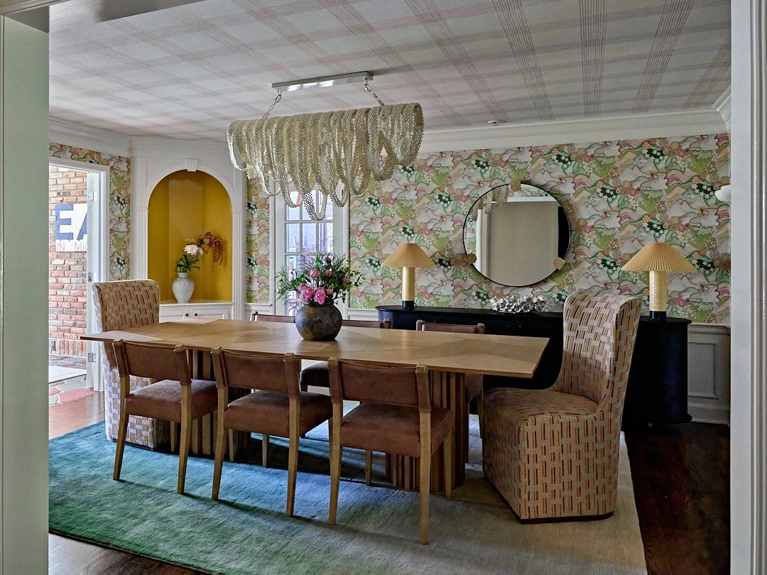 A dining room with a large wooden table, six chairs, and two patterned armchairs. The room features floral wallpaper, a large round mirror, a chandelier, and two matching lamps with yellow shades on a black sideboard. There is a vase with pink flower
