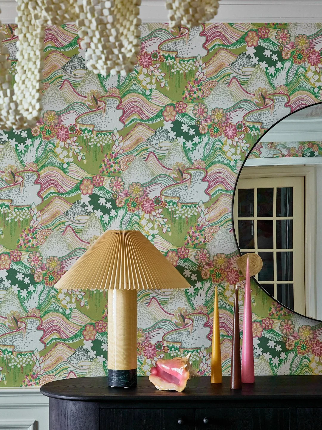 Decorative interior with a floral patterned wallpaper featuring pink, green, and white colors, a round mirror, and a black table holding a yellow pleated lampshade, colorful tall vases, and a pink shell ornament.