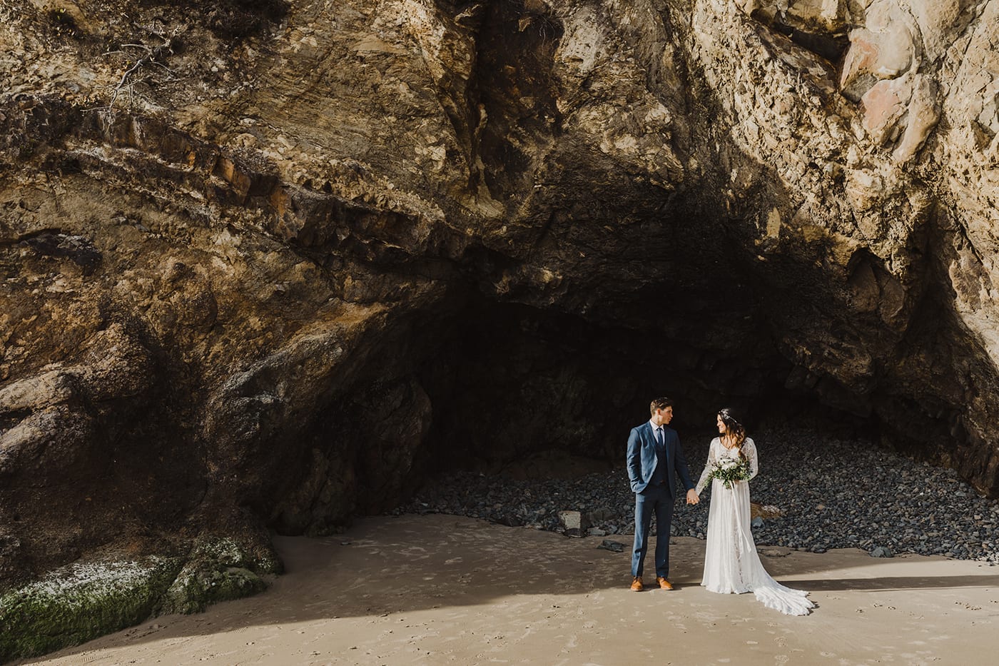 large rock formation with cave and couple standing in front holding hands before elopement