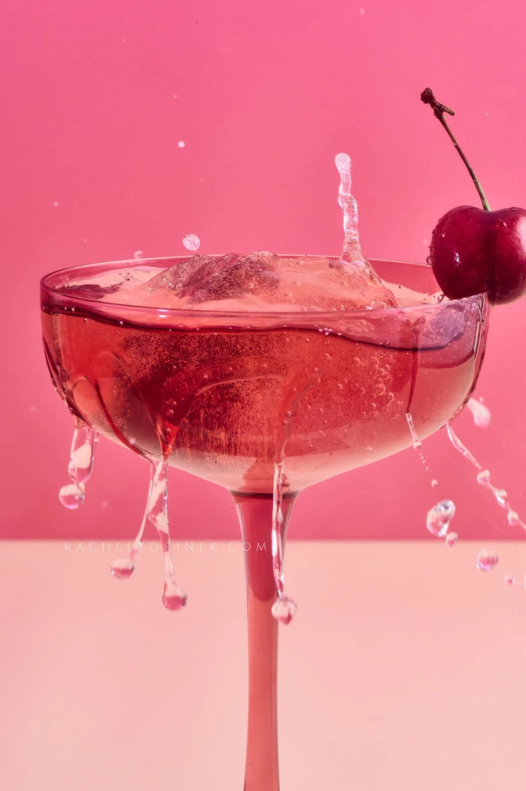  Pink drink in a pink glass with a cherry being dropped into the liquid. 