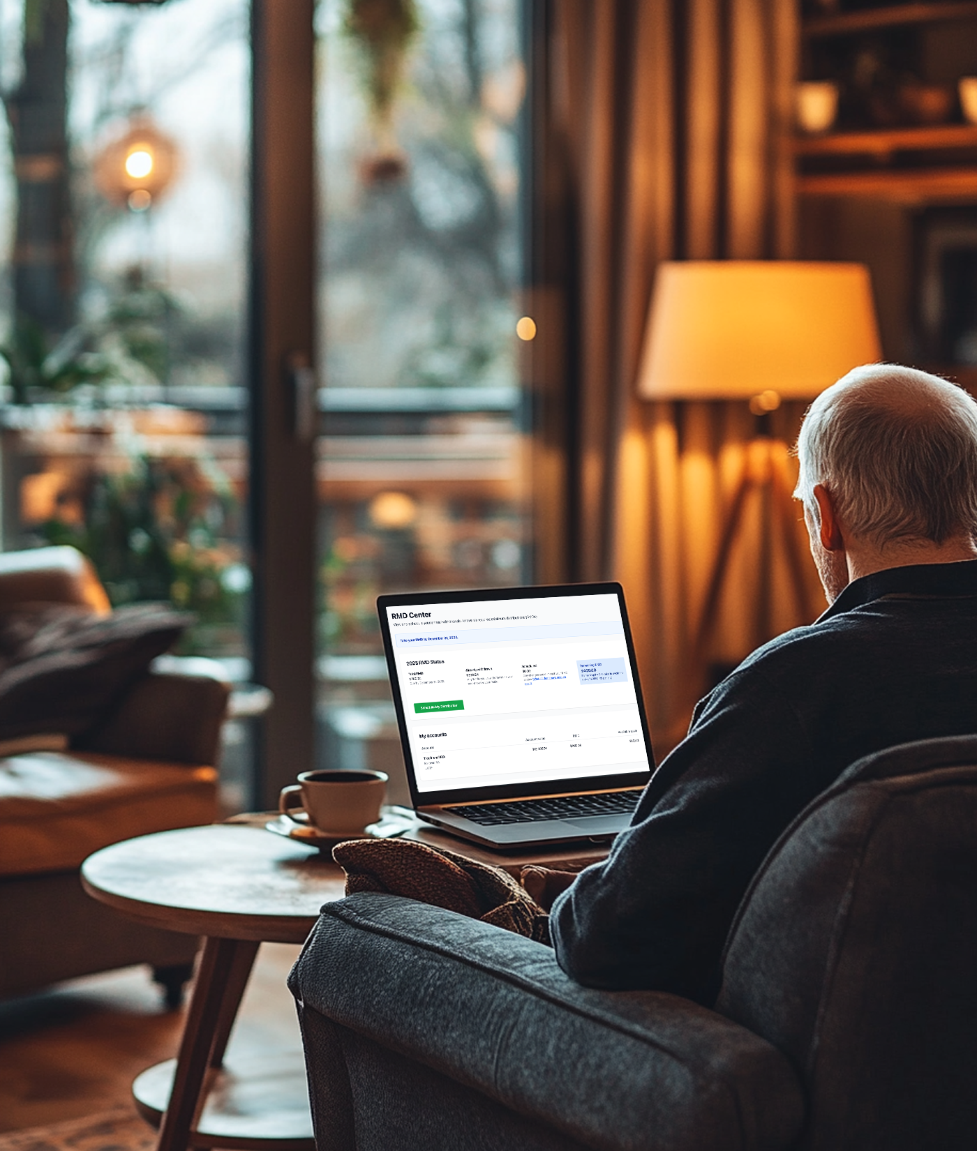 An elderly man sitting on a gray armchair, working on a laptop at a wooden side table in a cozy living room with warm lighting, a lamp, and large windows showing an outdoor view.