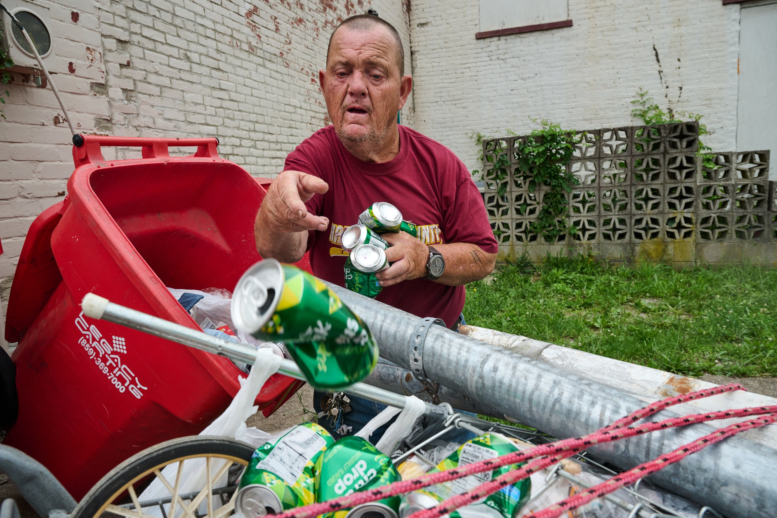  7/8/23—Cynthiana, Kentucky — Fryman tosses a can from a residential garbage can into his shopping cart. Fryman seeks permission from homeowners to look through their garbage cans for trash he can recycle, as there is no recycling infrastructure thro
