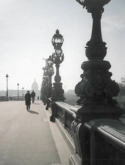 Paris in Fog: A few pedestrians cross a quiet bridge toward a domed building shrouded in mist, while elegant Parisian lampposts punctuate the scene. In black and white, this moment captures the city’s timeless, ethereal beauty.