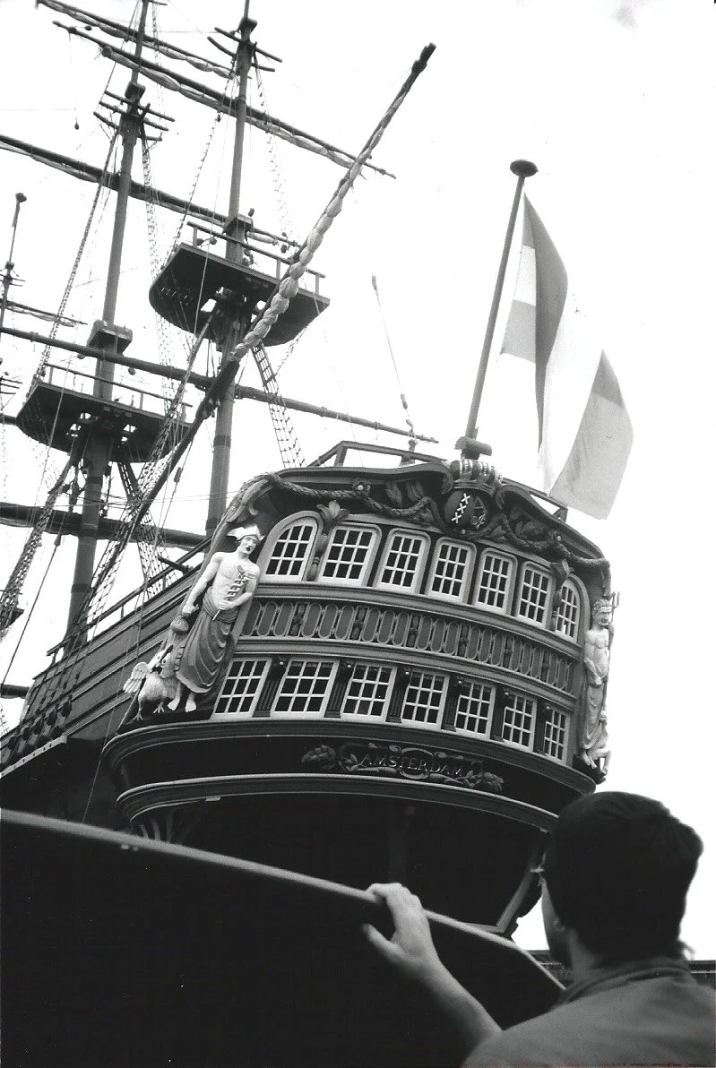 Amsterdam Harbor Awe: With his back to us, a man pauses at the railing, captivated by the massive ship before him. Its sculpted figurehead and towering masts evoke a quiet reverence—a fleeting moment of wonder on the water.