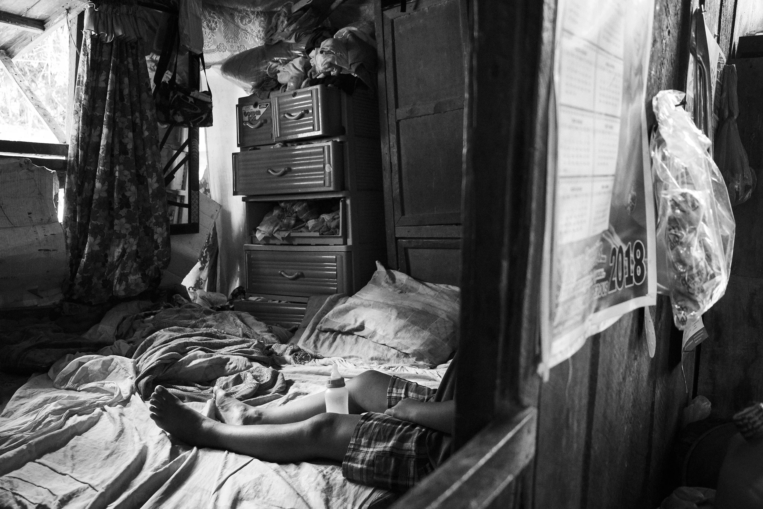 Black and white image of a cluttered bedroom with unmade bed, pillows, and a plastic drawer with clothes. A child's legs are visible on the bed, next to a baby bottle. A calendar is on the wall, dated 2018.