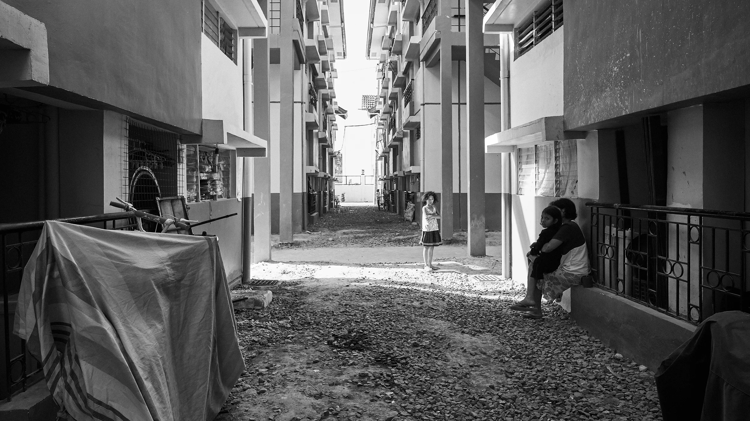 Black and white photo of a narrow alley between apartment buildings. A person stands in the middle, and two others sit on the right. A covered bicycle and rough gravel path are in the foreground.